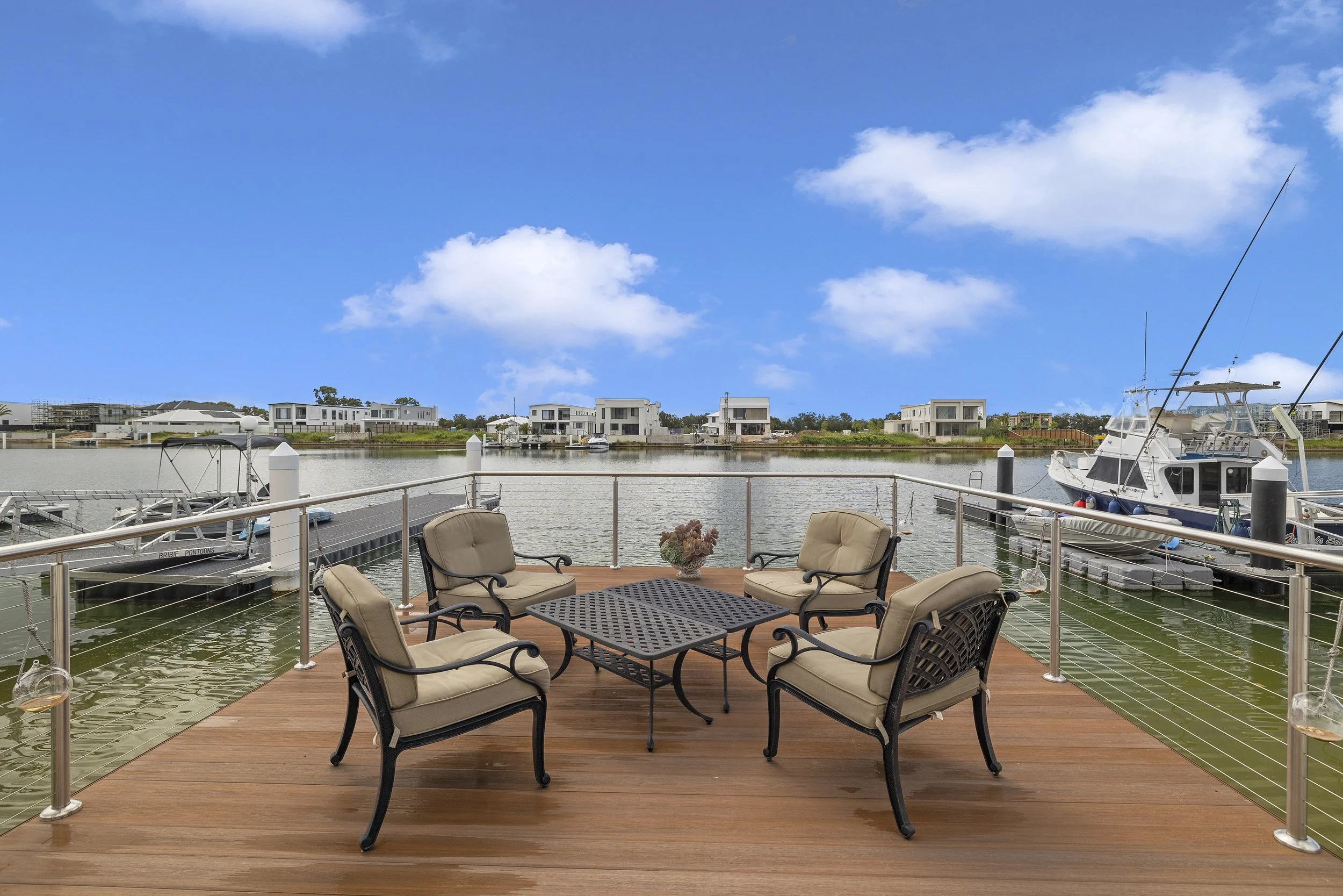 View of a waterfront patio with outdoor furniture, overlooking a marina with boats and modern houses in the background under a partly cloudy sky.