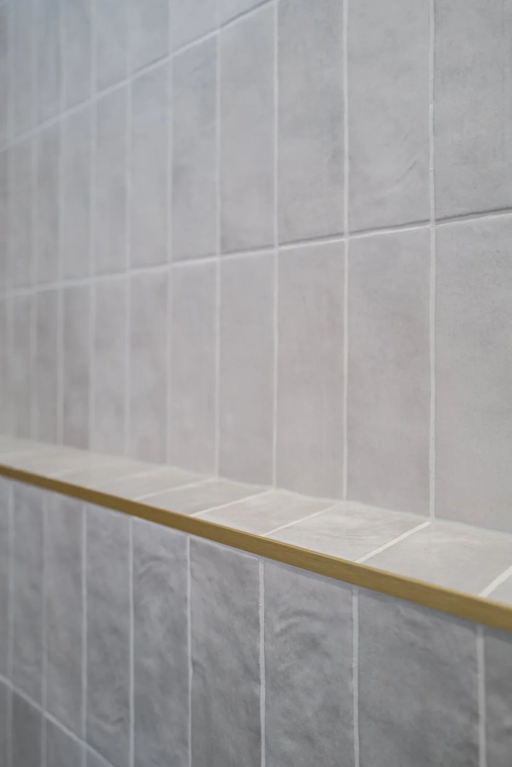 Close-up of light gray tiled wall with a narrow wooden shelf.
