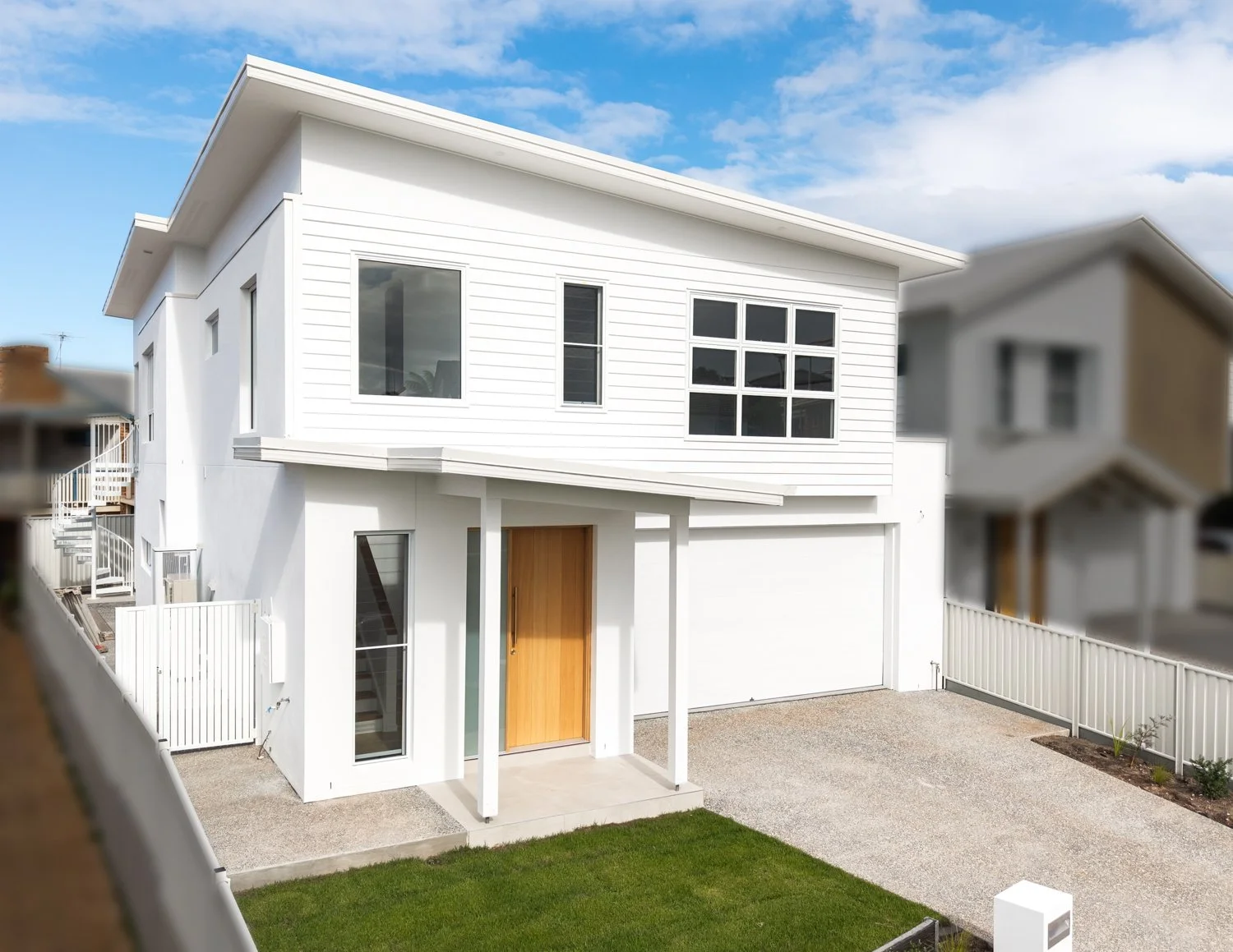 Modern two-story white house with wooden door, large front windows, and a small front yard with green grass, fenced yard, and a gravel driveway.