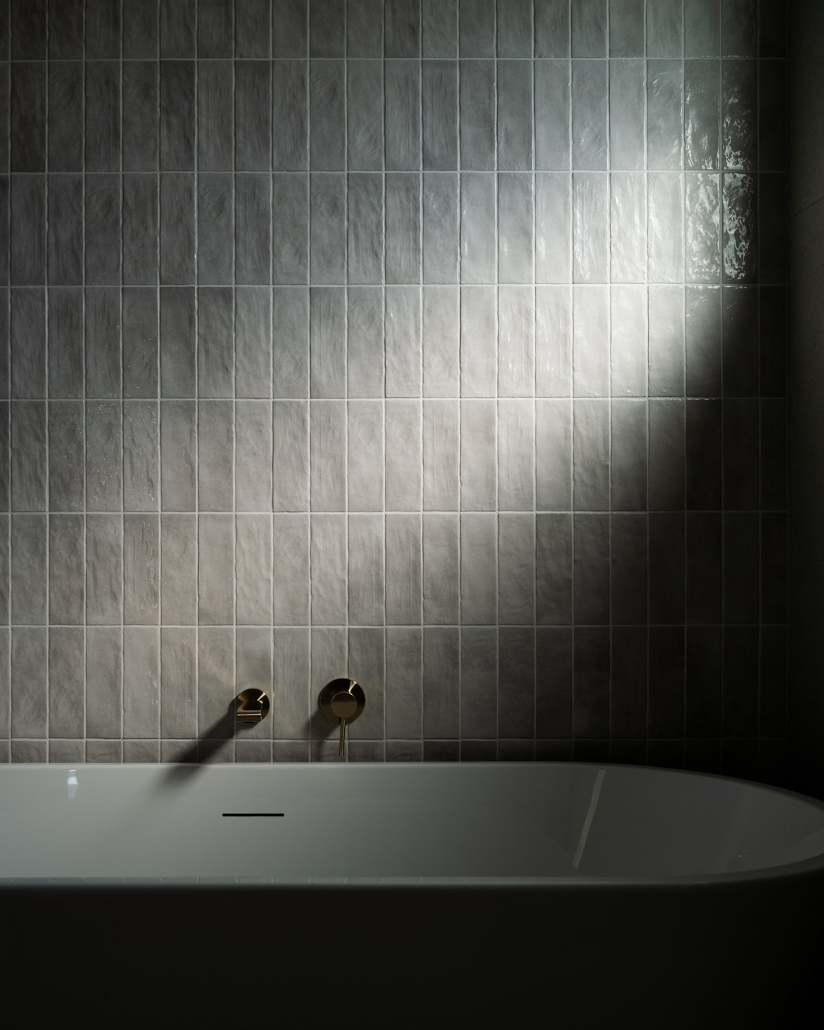 Shower with gray tiled wall, two gold-colored fixtures, and a white bathtub, illuminated by light reflecting off the tiles.