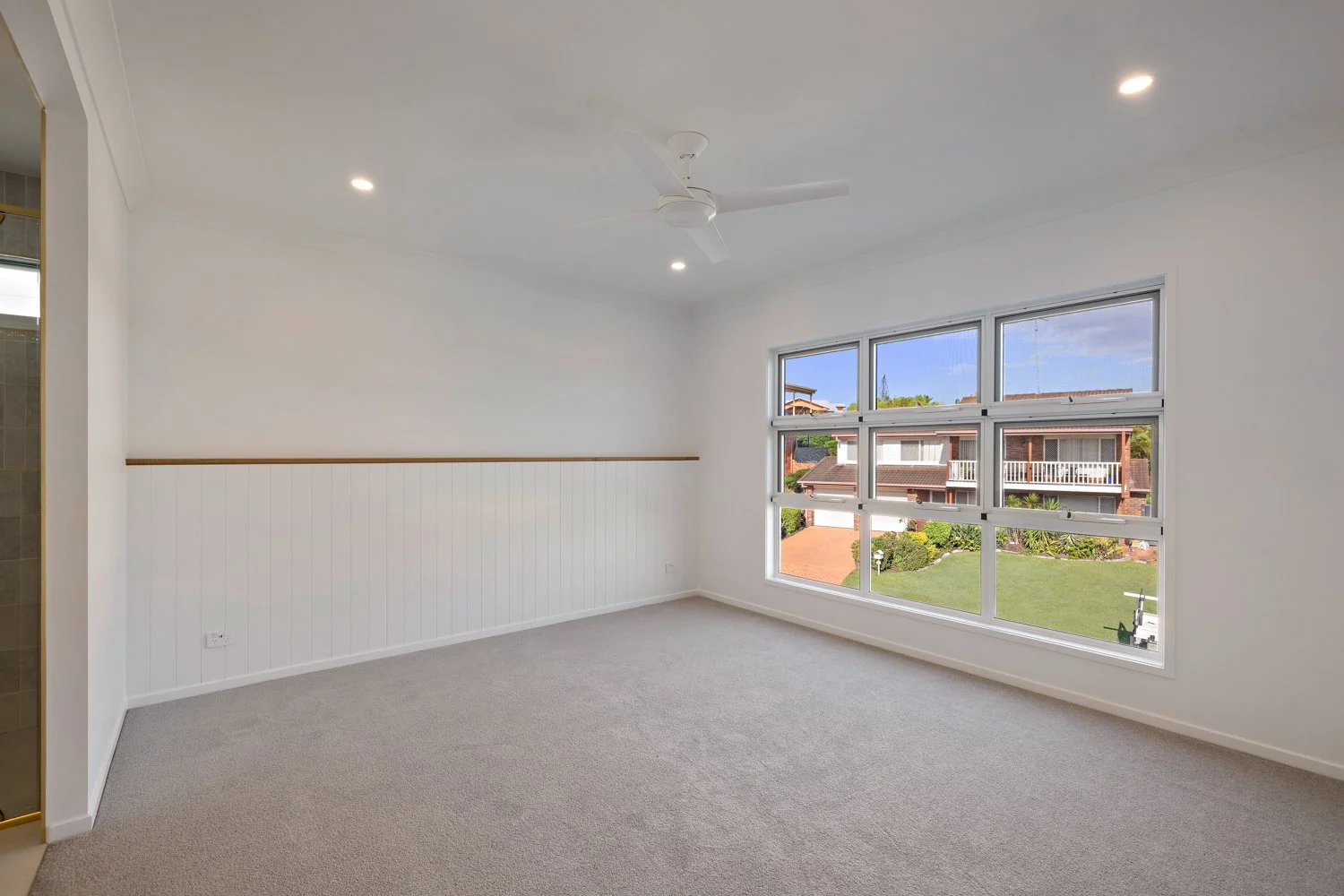 Empty room with white walls, beige carpet, large window, ceiling fan, and visible neighborhood outside.