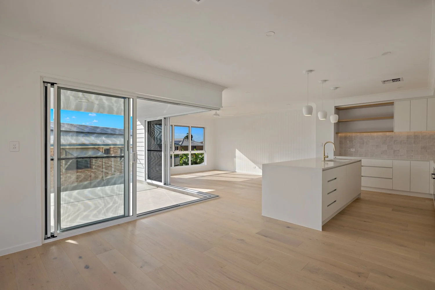 Empty modern white kitchen with an island, light wood flooring, and a patio with a sliding glass door leading outside.