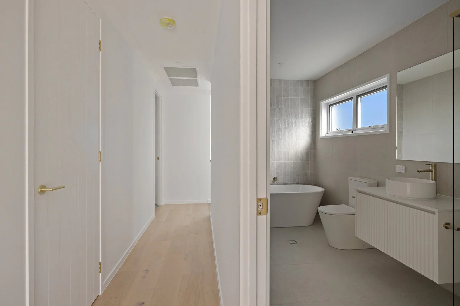Split view of a hallway leading to a bathroom with a bathtub, toilet, and vanity with a vessel sink, illuminated by natural light through a window.
