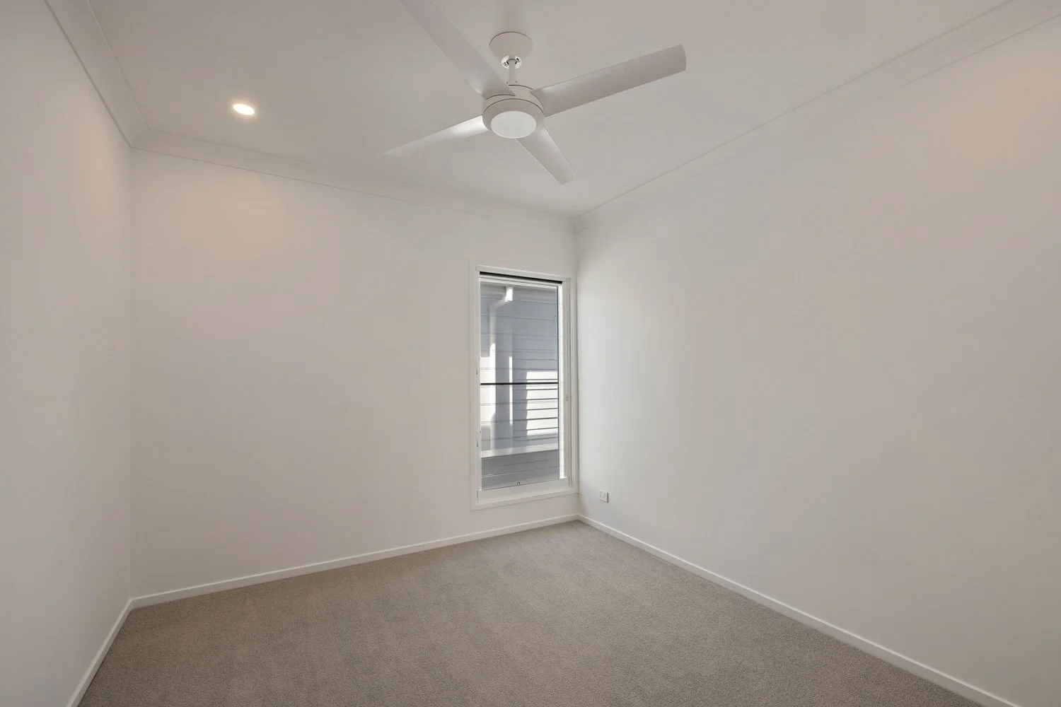 Empty white room with beige carpet, a ceiling fan, a single window with blinds, and a small balcony visible outside.