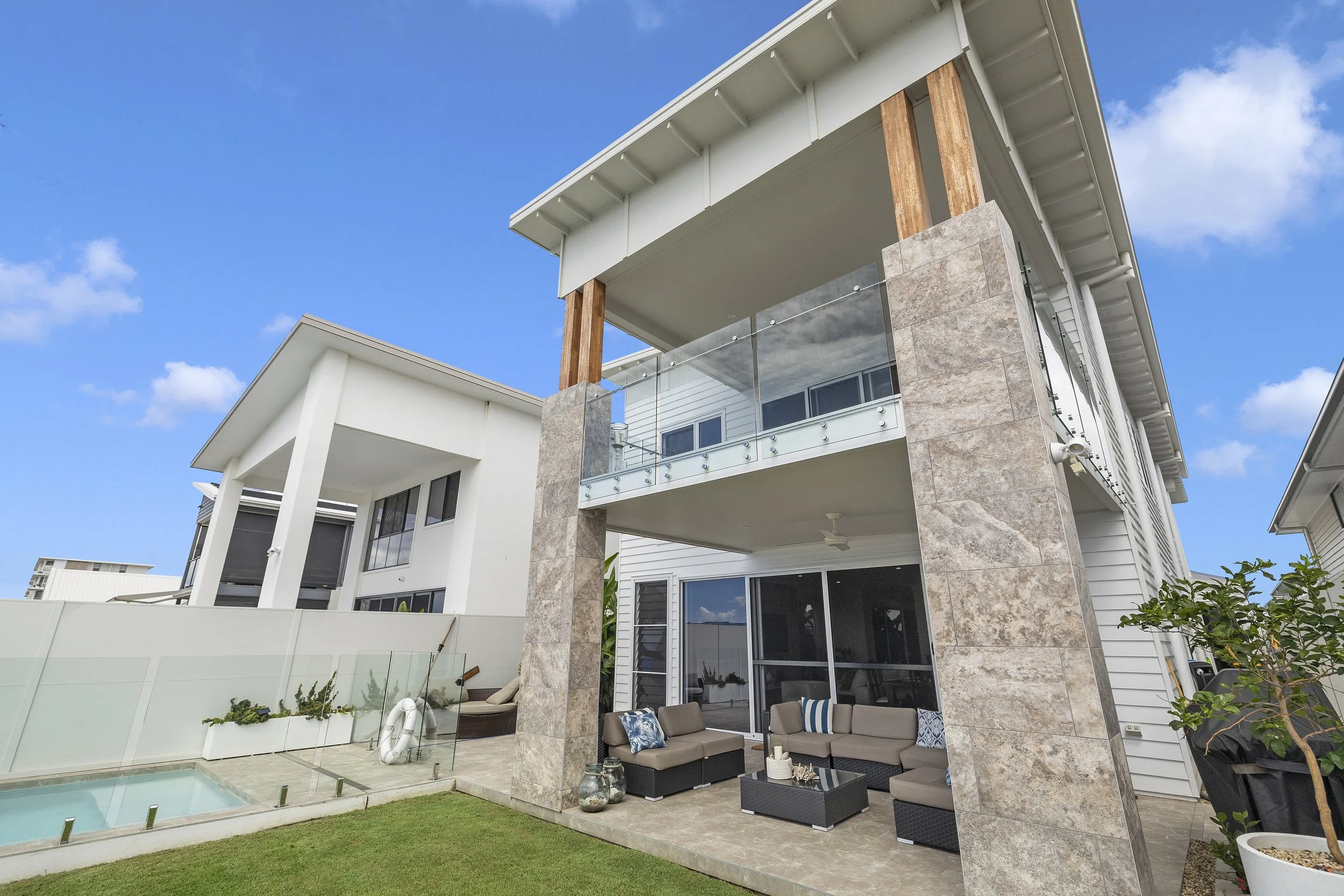 Modern multi-story house with outdoor patio area, glass railing, and pool, with blue sky and scattered clouds in the background.