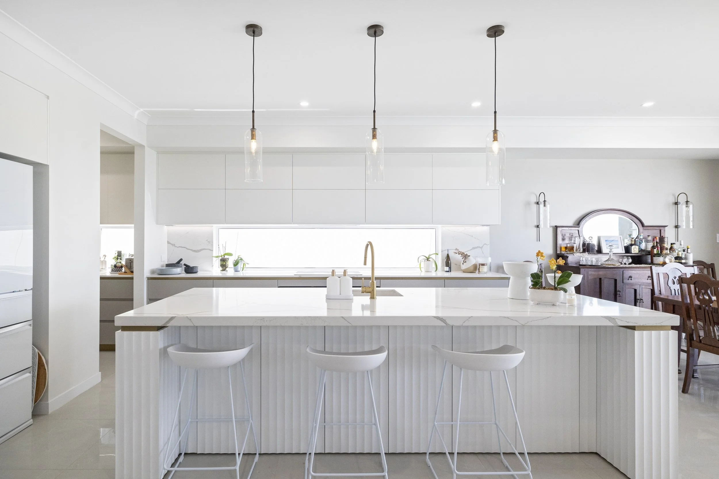 Modern white kitchen with a large island, three white bar stools, and pendant lighting hanging from the ceiling.