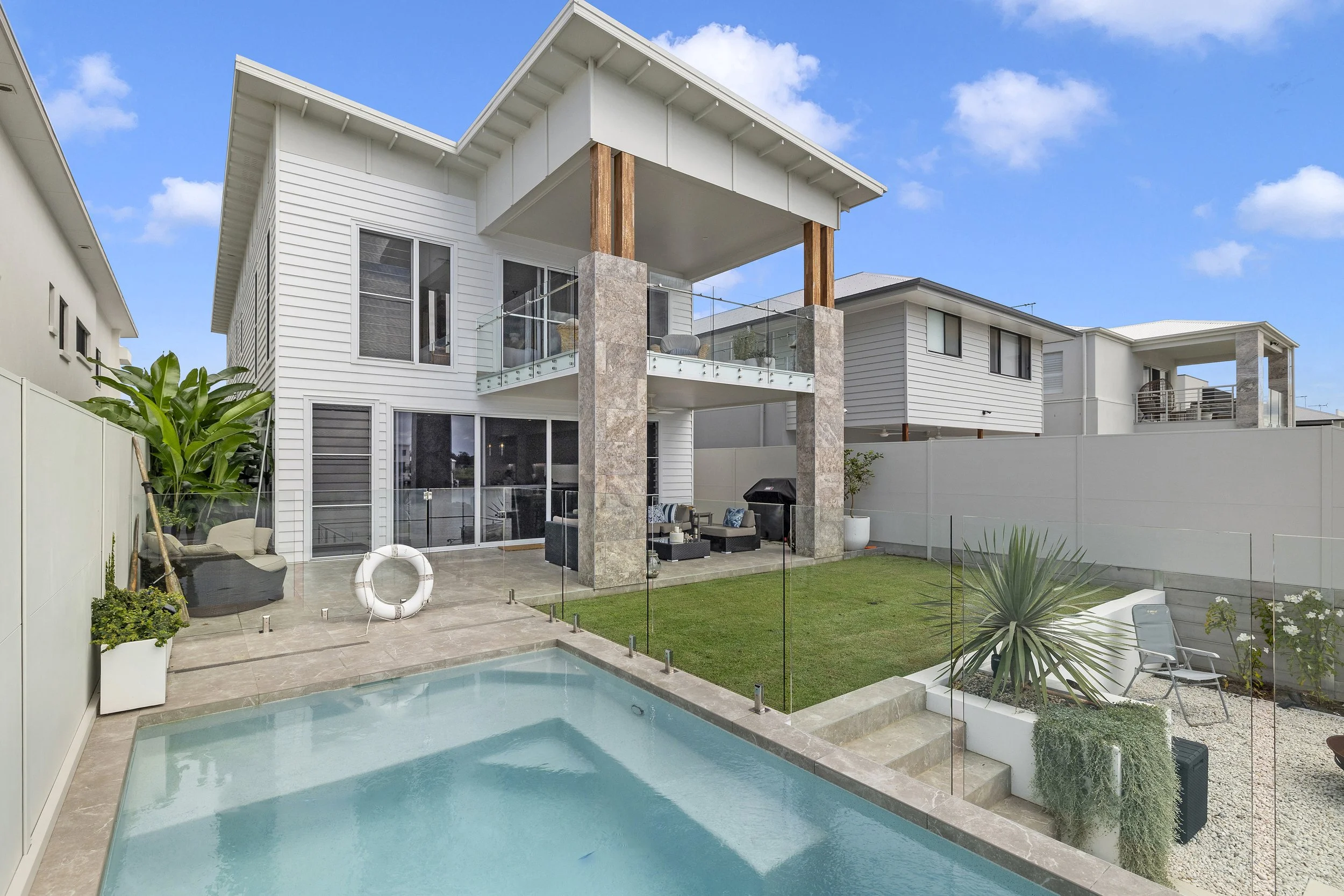 Modern backyard with a small swimming pool, outdoor lounge area with chairs and a grill, enclosed by a glass fence, with a white multi-story house featuring an upper balcony in the background and a blue sky with clouds.