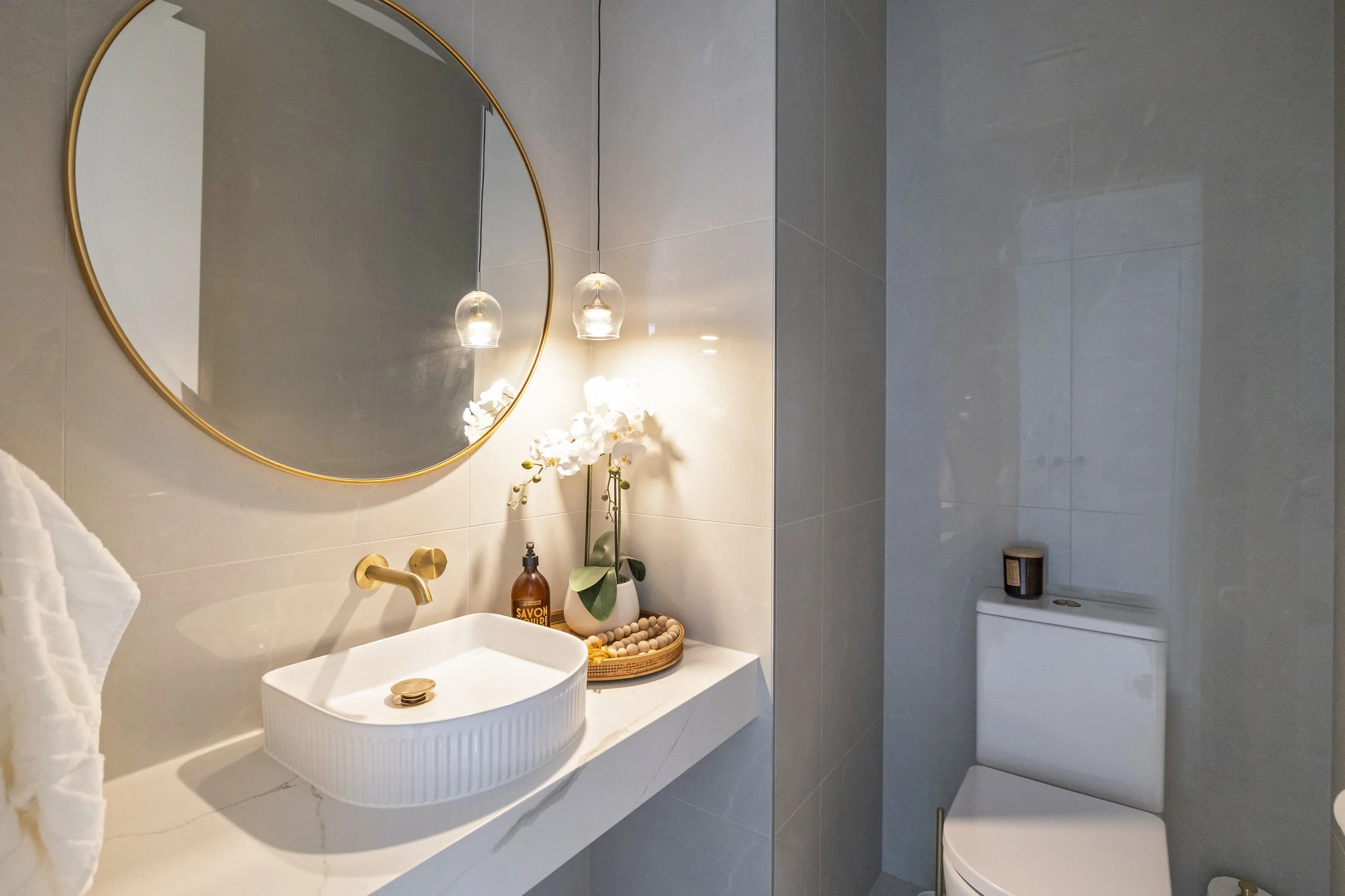 Modern bathroom with a white sink, a large round mirror with a gold frame, and two hanging pendant lights. There is a vase with white orchids on the counter, along with soap and a basket of wooden balls. To the right, a toilet and a small container a
