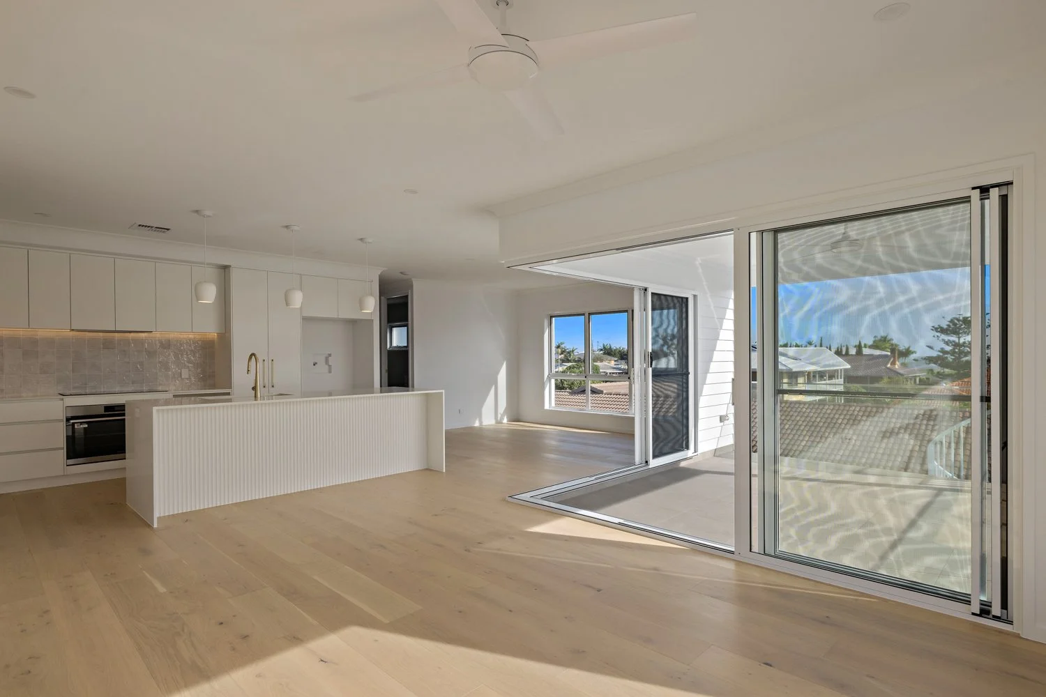 Empty modern living space with a white kitchen and large sliding glass doors opening to a balcony, view of rooftops and blue sky