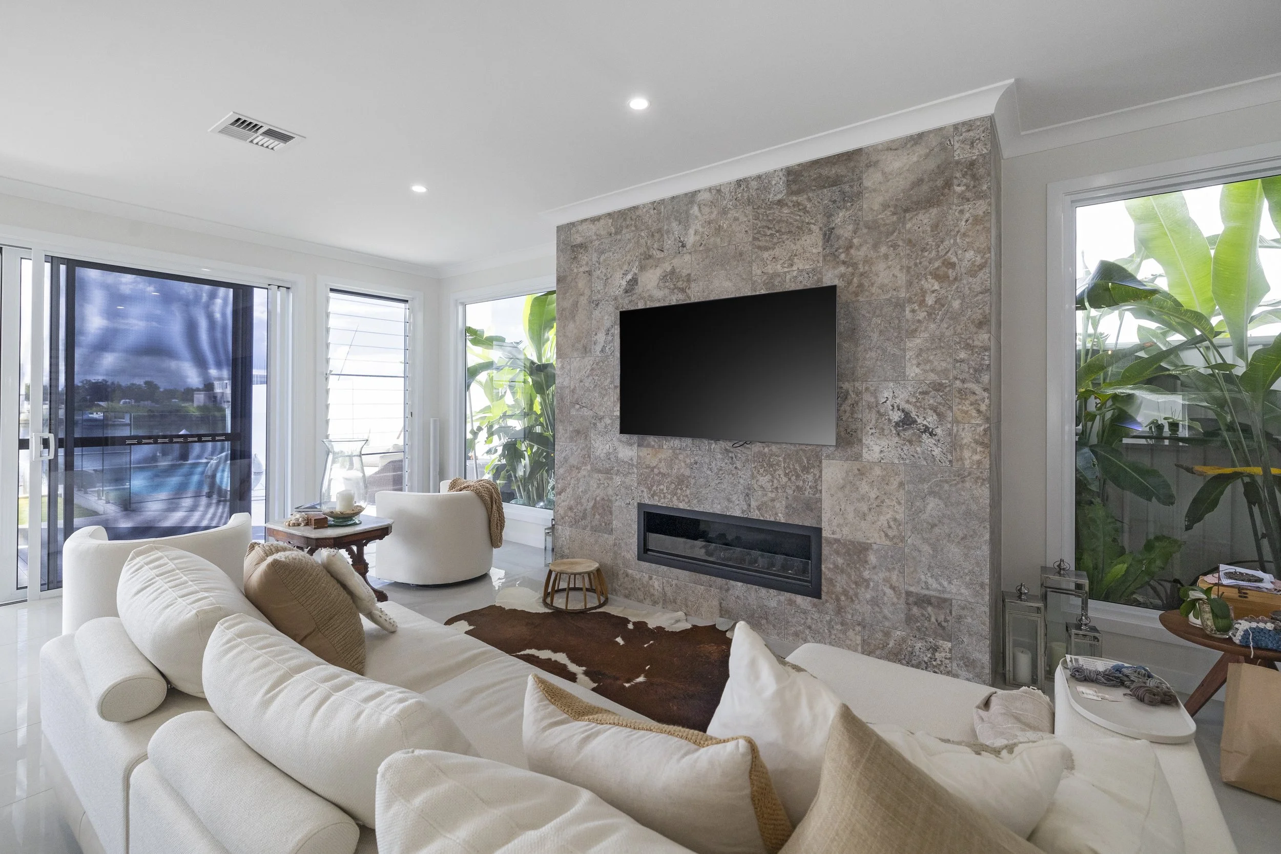 Living room with white couches, a stone fireplace with a mounted flat-screen TV, large windows with blinds, and leafy plants outside, bright and airy with natural light.