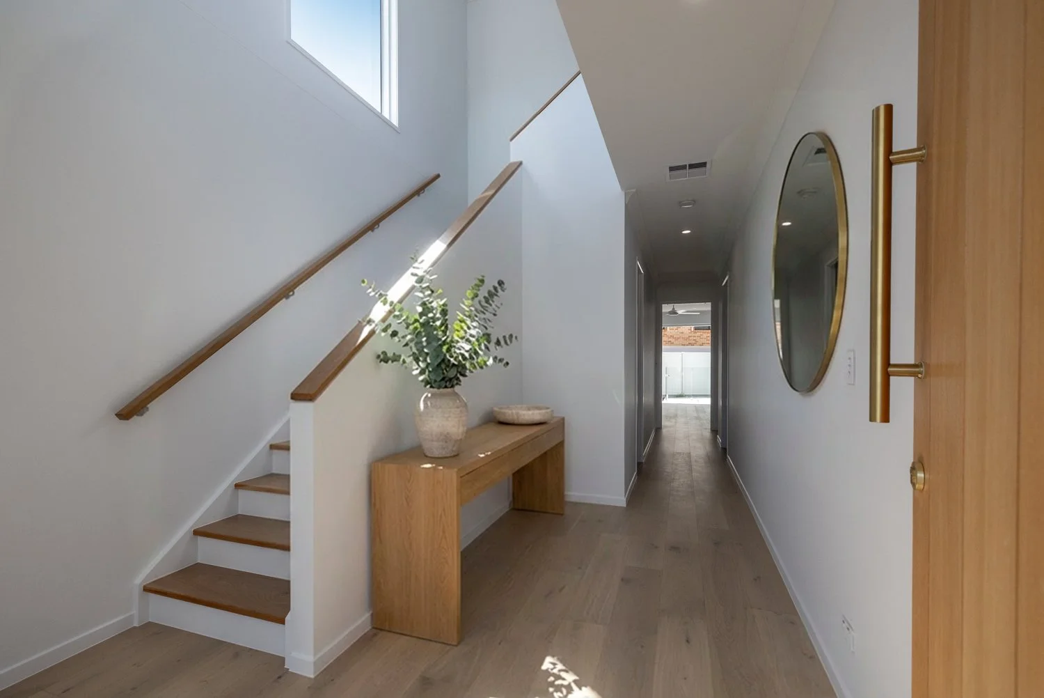 Modern interior hallway featuring a staircase with wooden steps and railing, a wooden console table with decorative vase and bowl, round wall mirror with gold frame, and a wooden door on the right side.