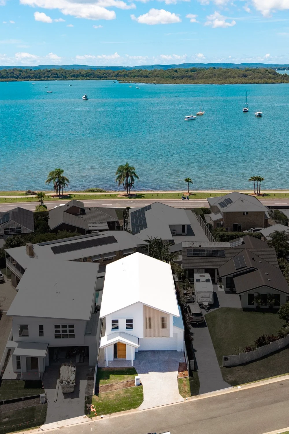 Aerial view of a neighborhood near a large blue body of water with boats, a road, palm trees, and houses, including a modern white house at the forefront.