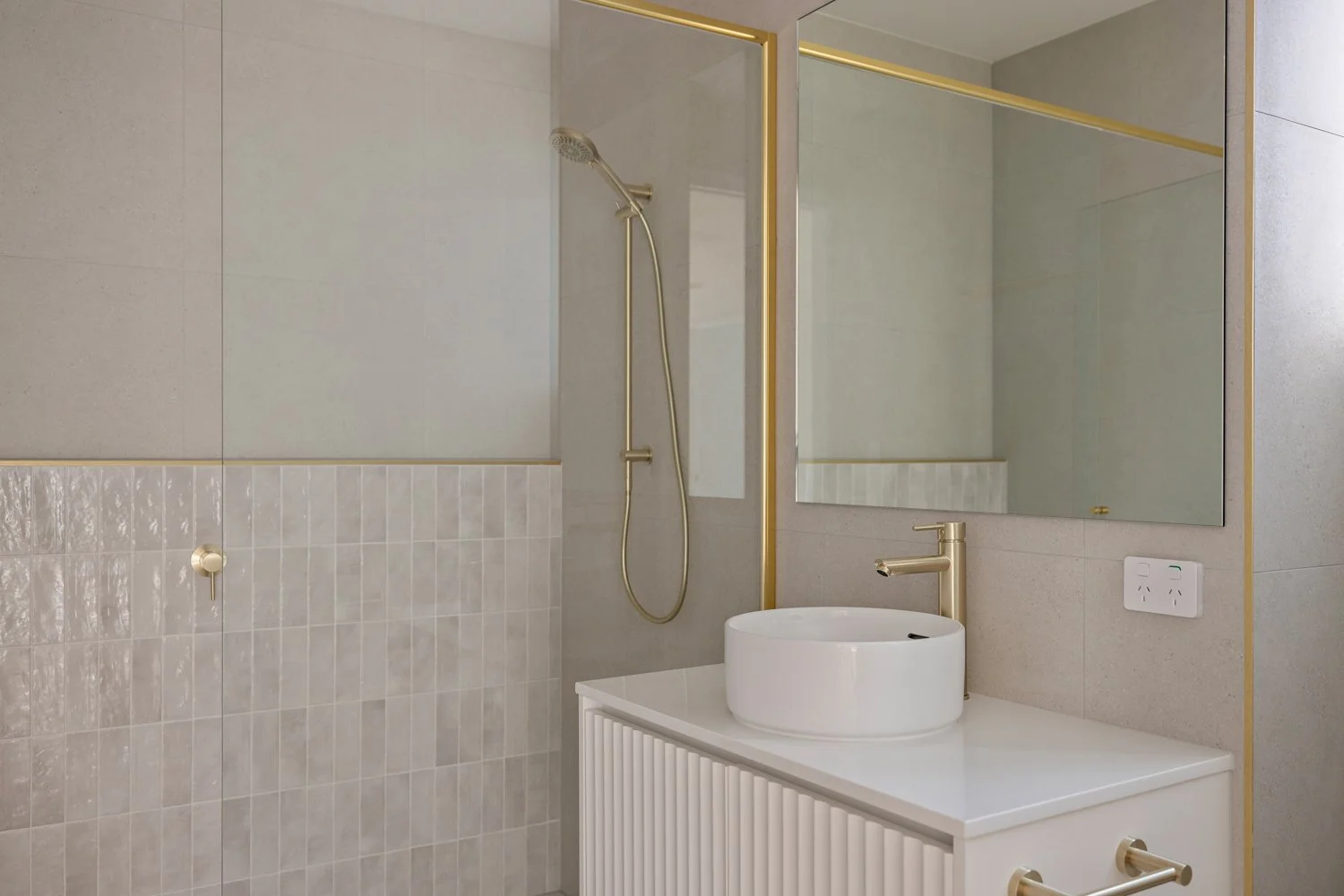 Modern bathroom with beige tiles, a round white vessel sink, gold fixtures, a large mirror, and a glass shower enclosure with a gold frame.