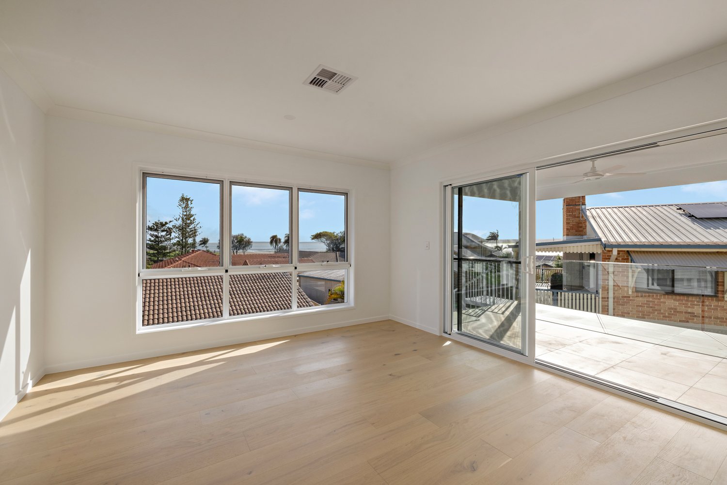Empty room with large windows and sliding glass door, wooden floor, overlooks neighboring rooftops and trees