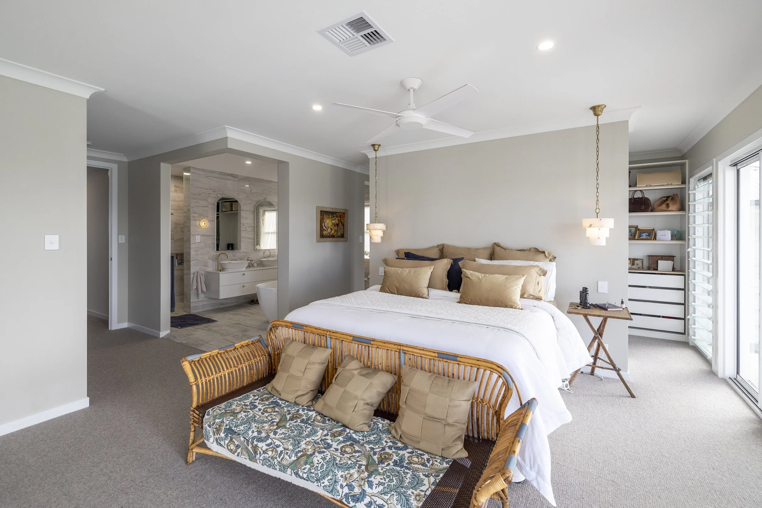 Interior of a modern bedroom with a bed, wicker sitting area, and an open view to a bathroom. The room has white walls, a ceiling fan, hanging pendant lights, and large windows with shutters.