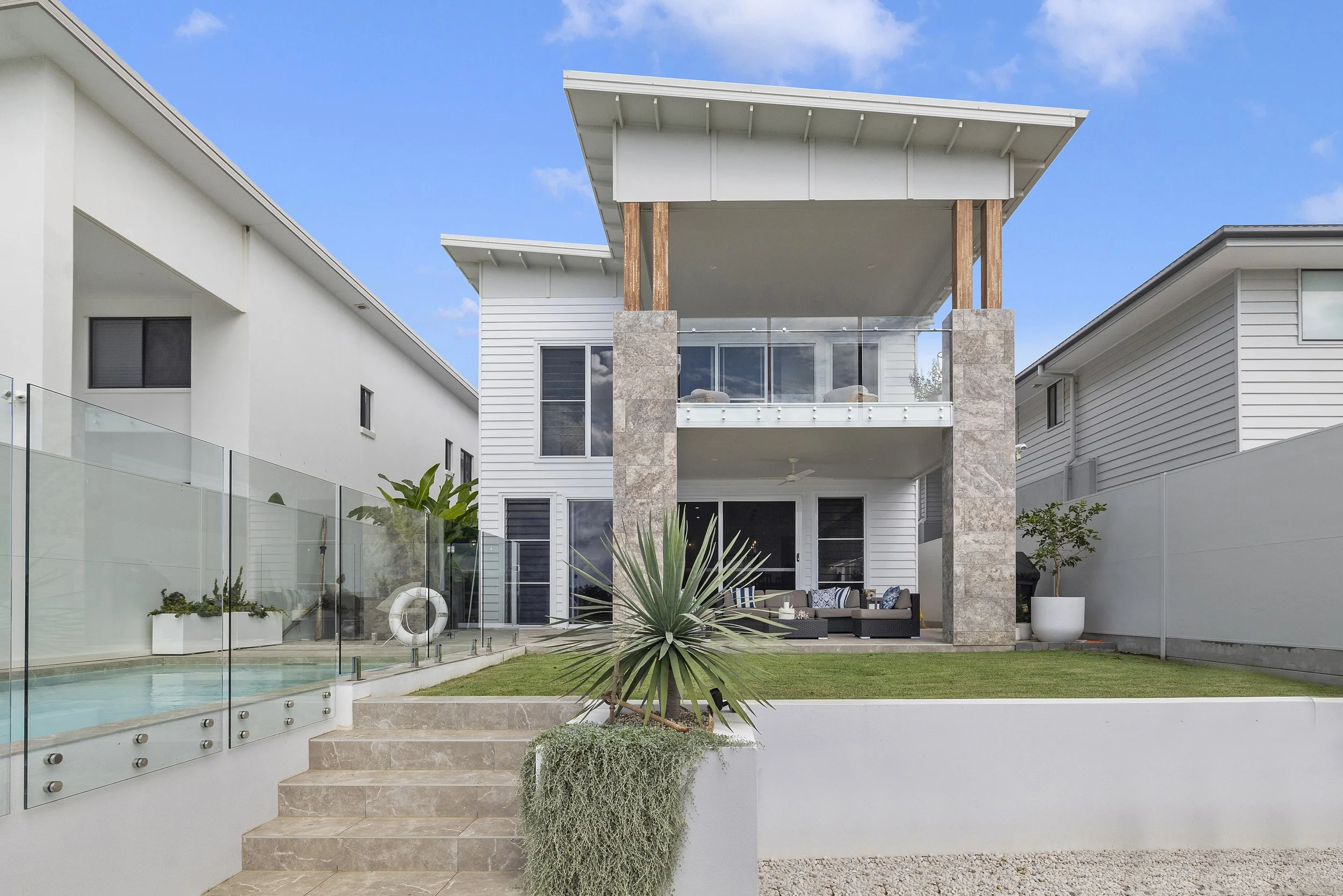 Modern multi-story house with white exterior, large windows, a balcony with glass railing, and a spacious backyard featuring a grilling station, outdoor seating, plants, and a small pool.