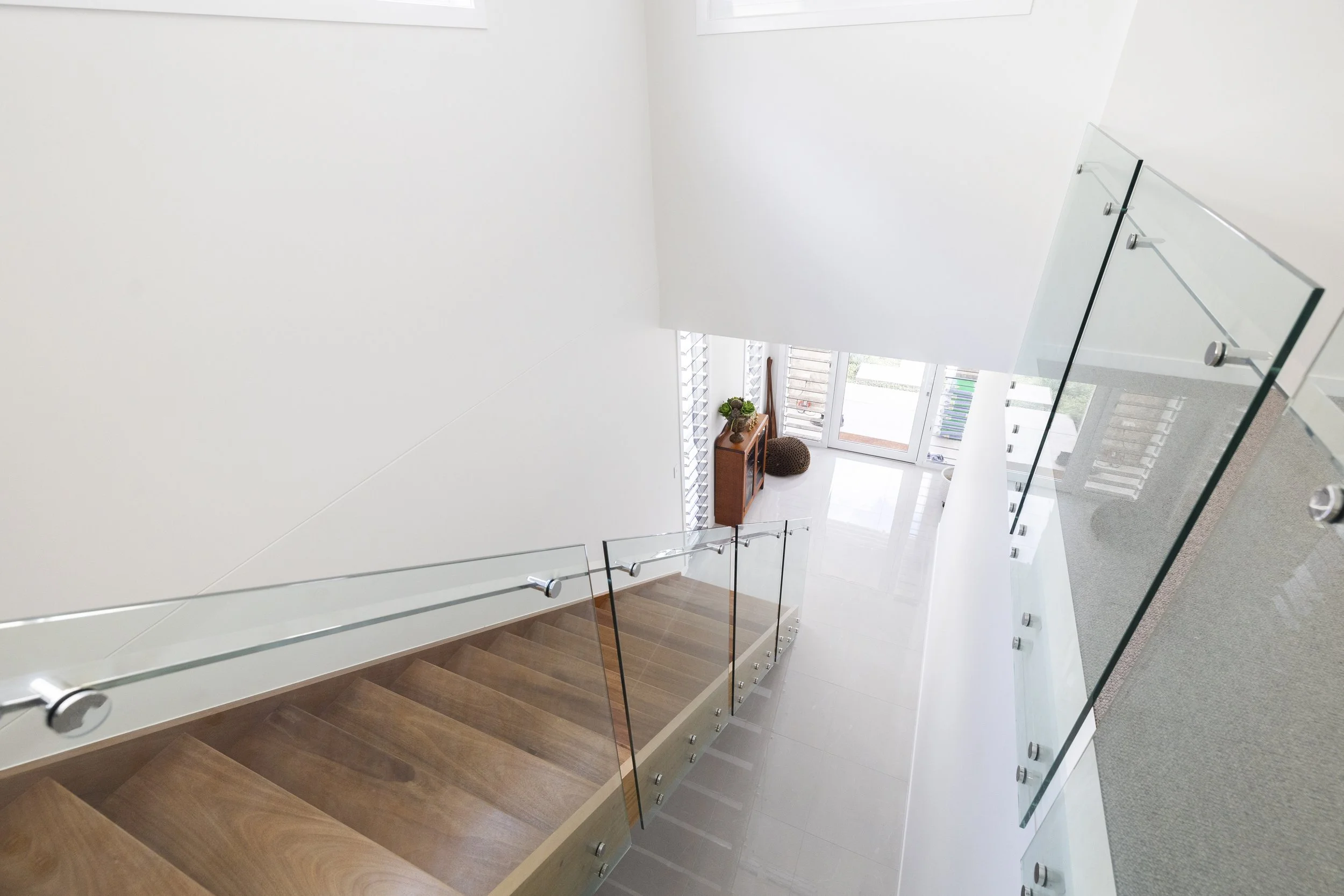 Upward view of a staircase with wooden steps, glass railing, and metal fixtures, leading to a bright area with a white wall, window with blinds, and a small table with decorative items.