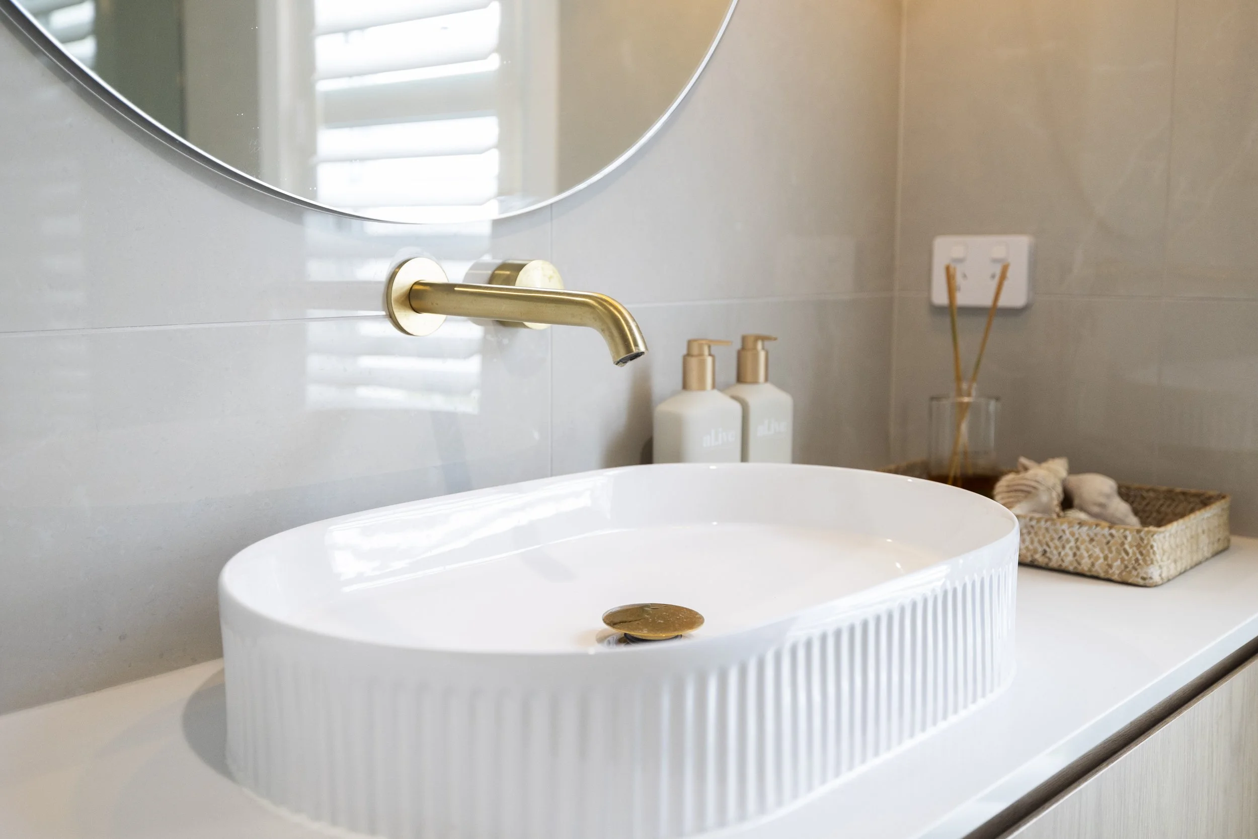 Bathroom sink with a round white basin, a gold faucet, two white soap dispensers labeled 'olive,' a small glass container with reed diffusers, a wicker basket with seashells, and a mirror with a window reflected in it.