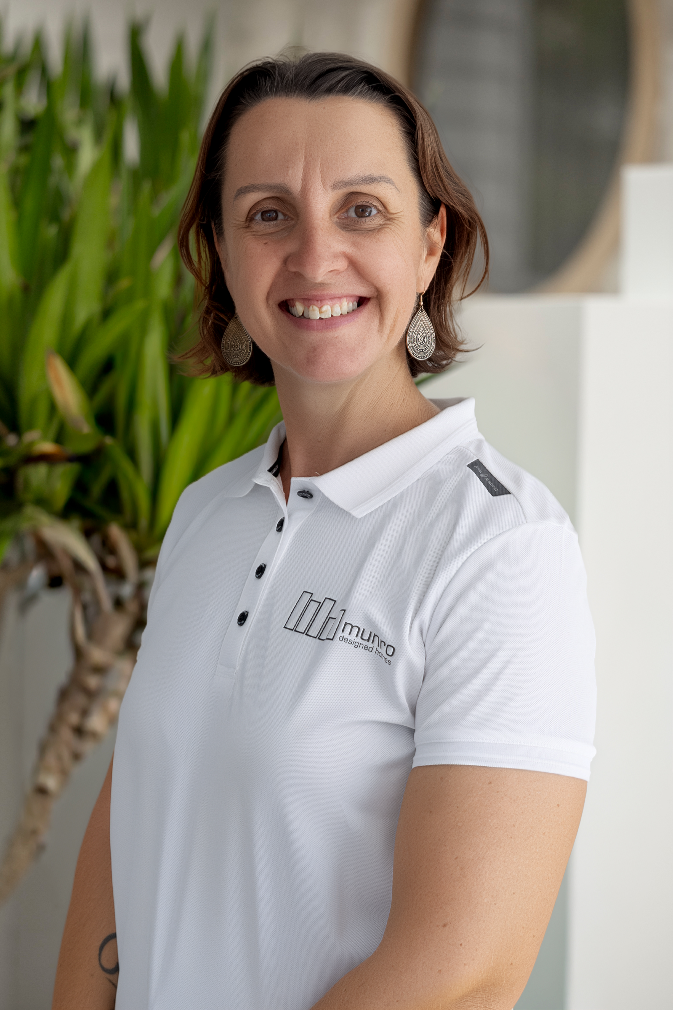 A woman with short dark hair smiling, wearing a white polo shirt and large dangling earrings, standing indoors with a green plant and decorative wall element in the background.