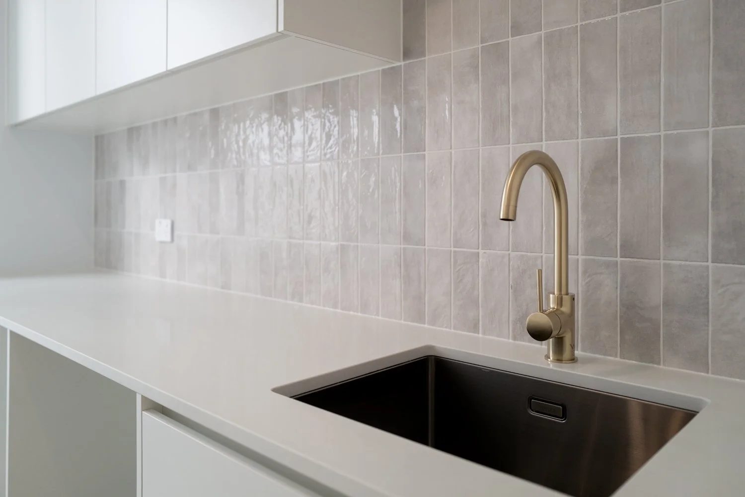 Modern kitchen countertop with a black sink and brushed gold faucet, beige tiled backsplash, and white cabinetry.