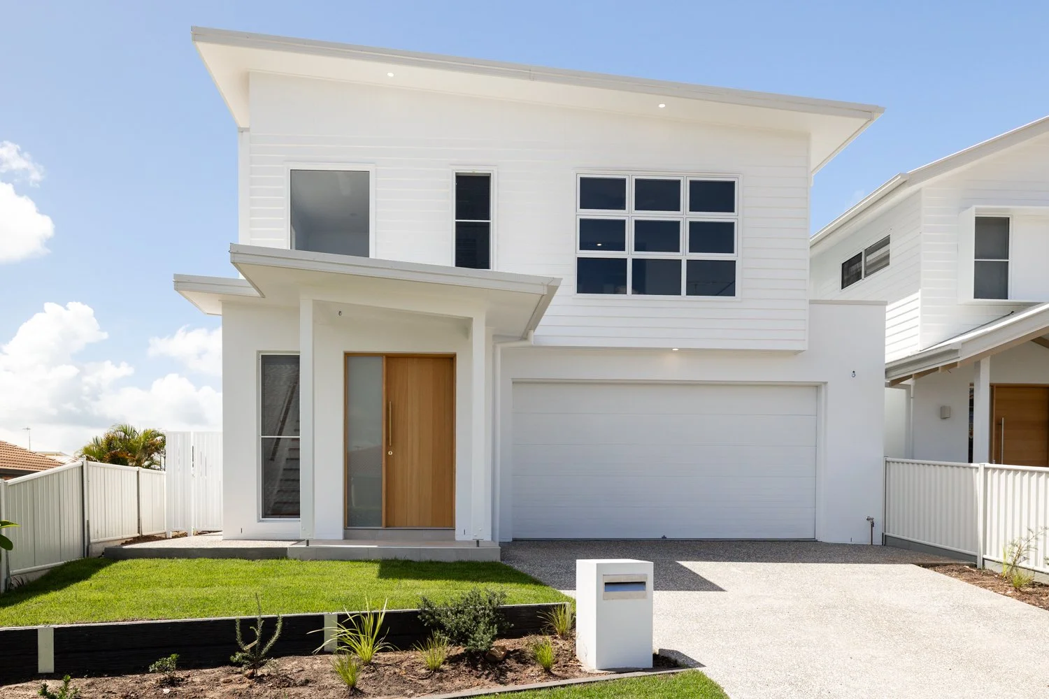 Modern white two-story house with a wooden front door, large windows, and a garage, surrounded by a small garden with grass and plants, under a partly cloudy sky.