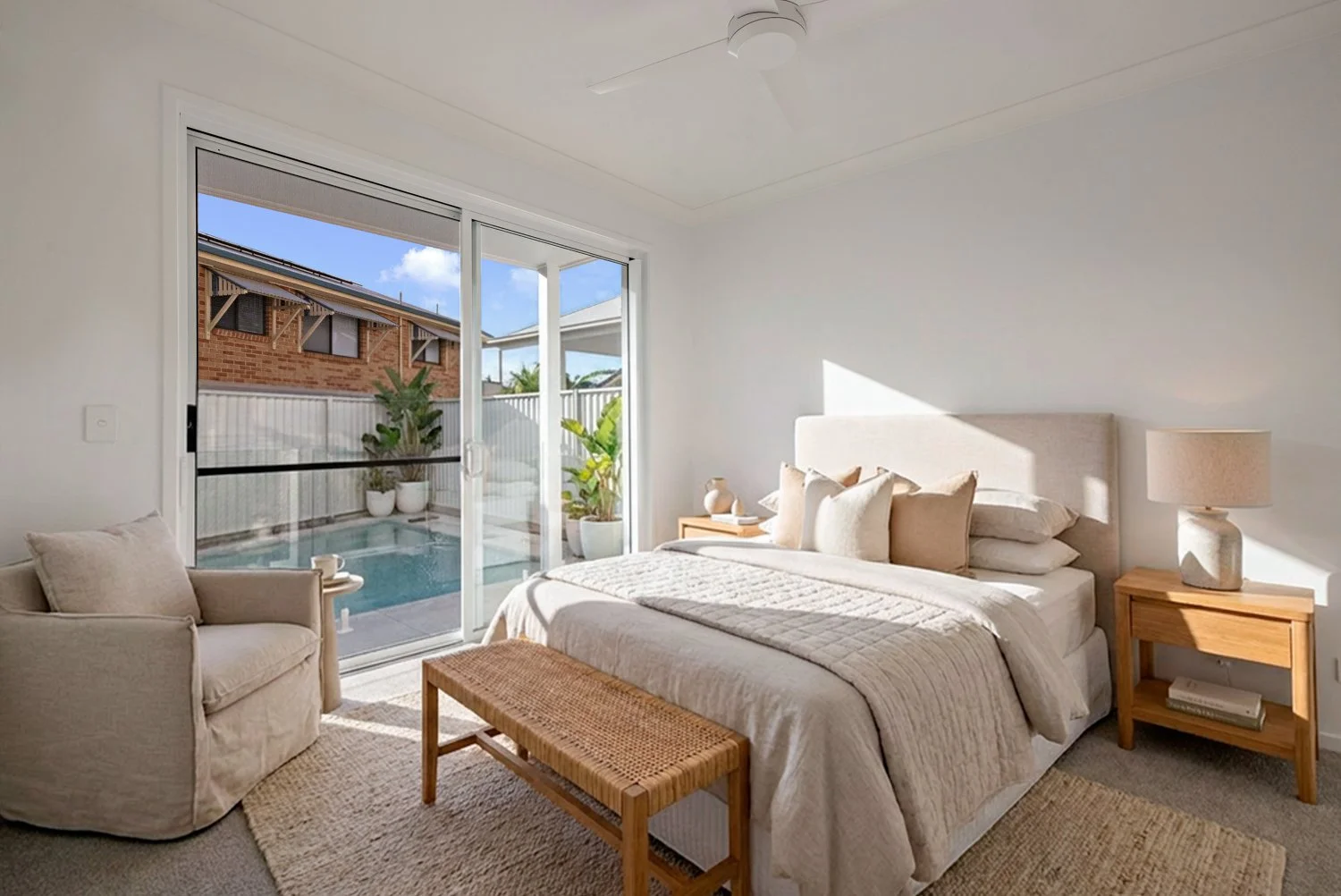 Bright bedroom with white bed, beige pillows, matching bedside tables, a lamp, a woven bench, and a sliding glass door leading to a small outdoor patio with potted plants and a pool.