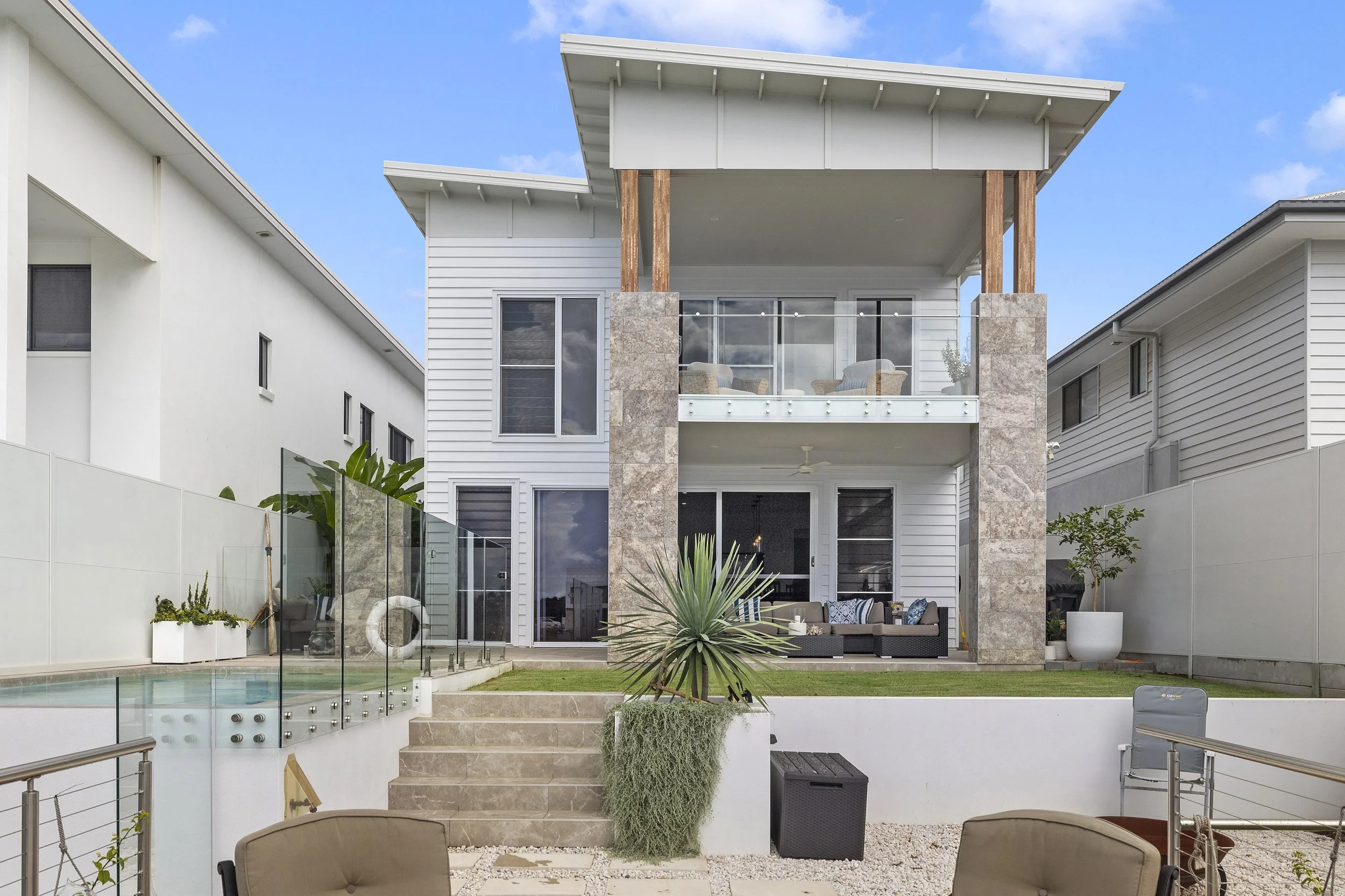 Exterior view of a modern two-story house with a balcony, large windows, and a backyard pool with a glass fence. There are outdoor furniture and potted plants.