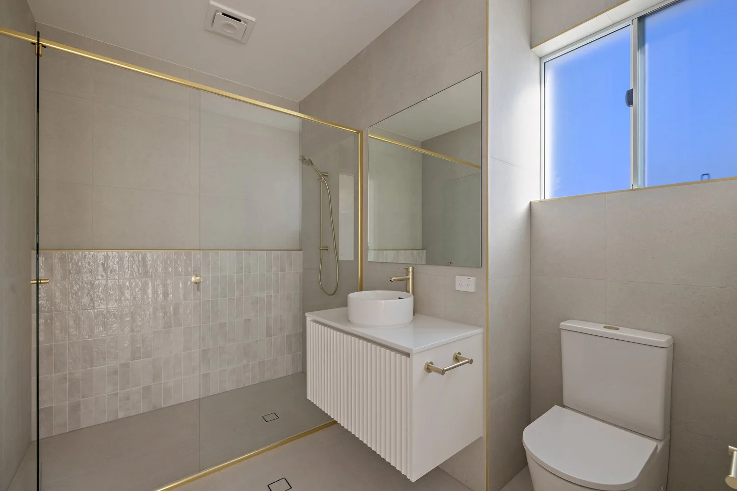 Modern bathroom featuring a walk-in shower with glass walls, a white sink vanity with a round vessel sink, a gold faucet, a large mirror, a toilet, and a window with blue sky visible outside.