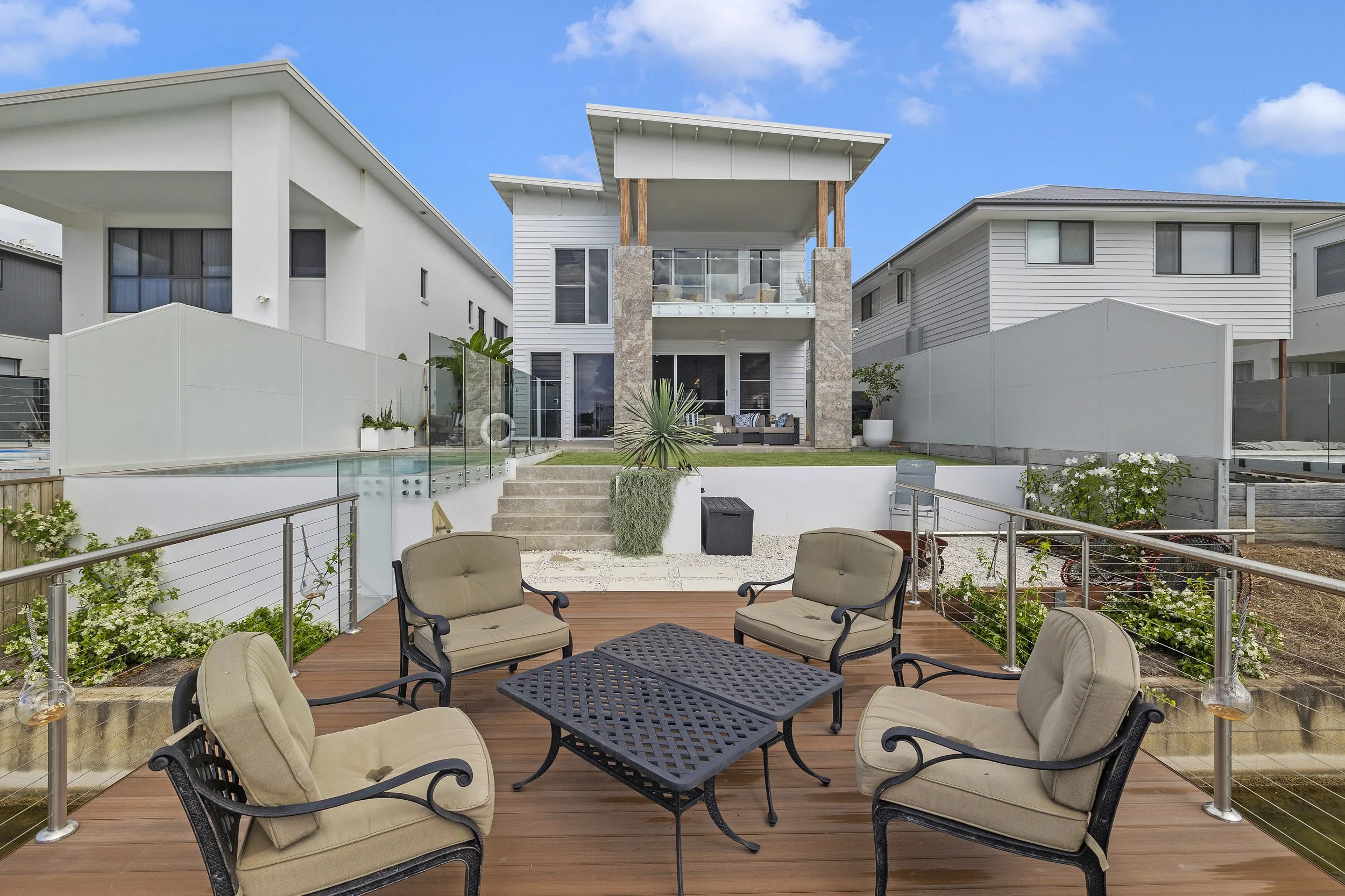 A backyard patio with outdoor seating, a pool with glass fencing, and modern houses in the background under a blue sky.