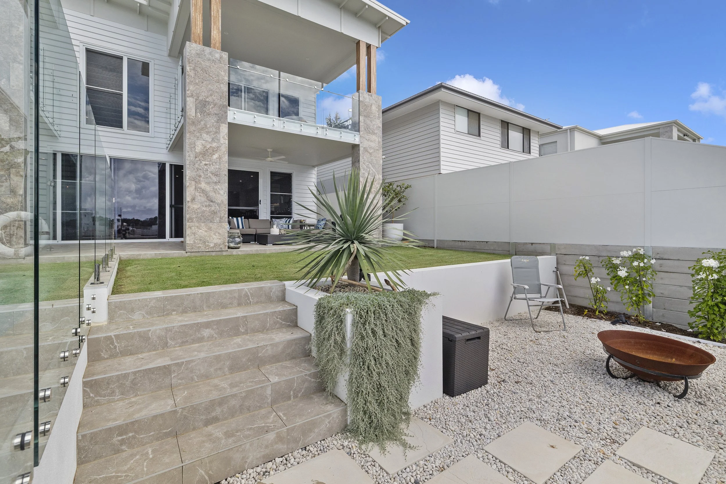 Modern backyard with stone steps leading to a patio with outdoor furniture, a small garden area with a palm plant, and a high white fence.