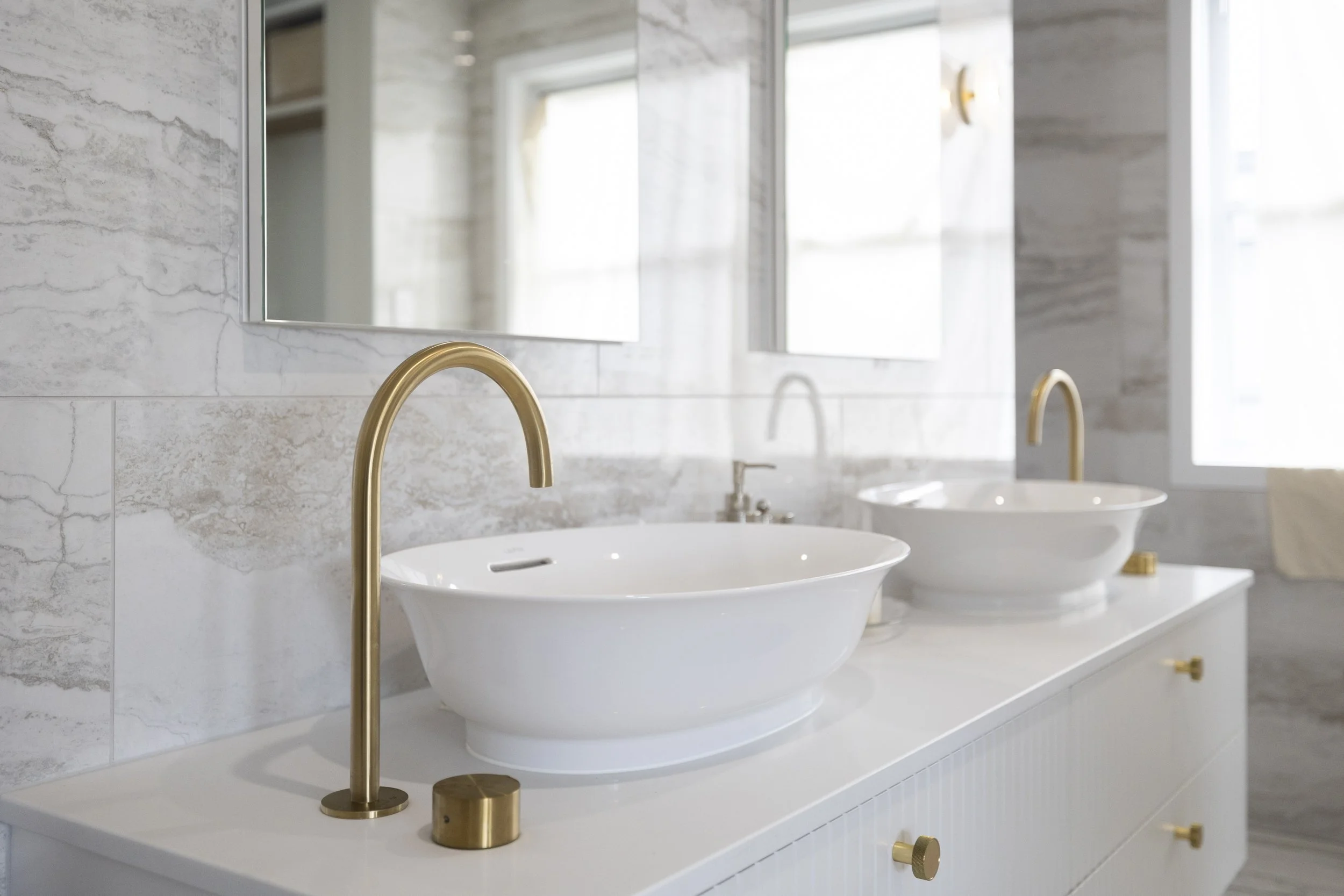 A modern bathroom with a white marble countertop holding two white vessel sinks with gold faucets and knobs, a large mirror, and natural light coming in through windows.