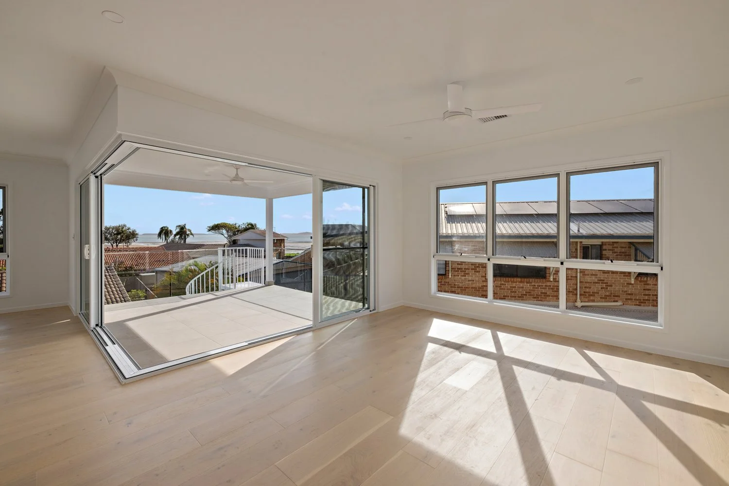 Empty living room with large windows and glass sliding door leading to a balcony with a view of neighboring houses and trees.