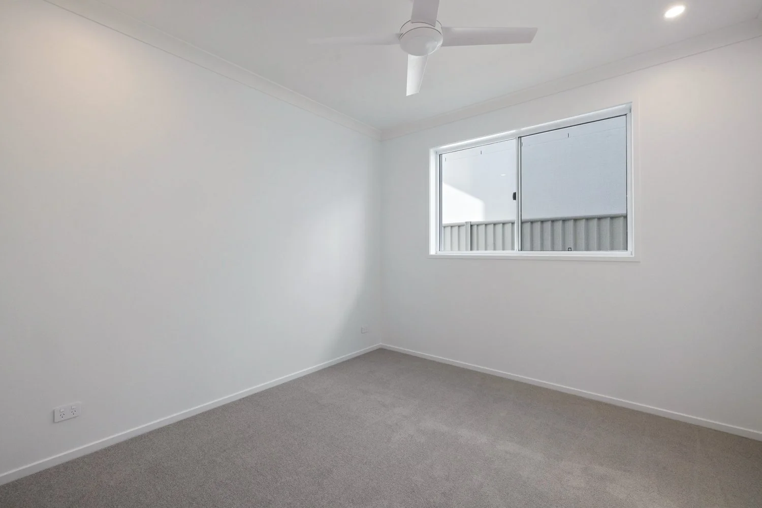 Empty white room with a window, ceiling fan, beige carpet, and electrical outlet.