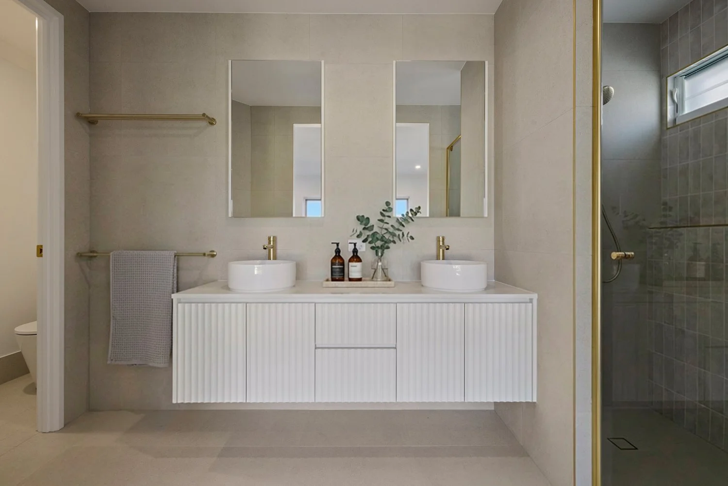 Modern bathroom with a double vanity featuring two vessel sinks, gold fixtures, and a large mirror, with a shower on the right and a toilet partially visible on the left.