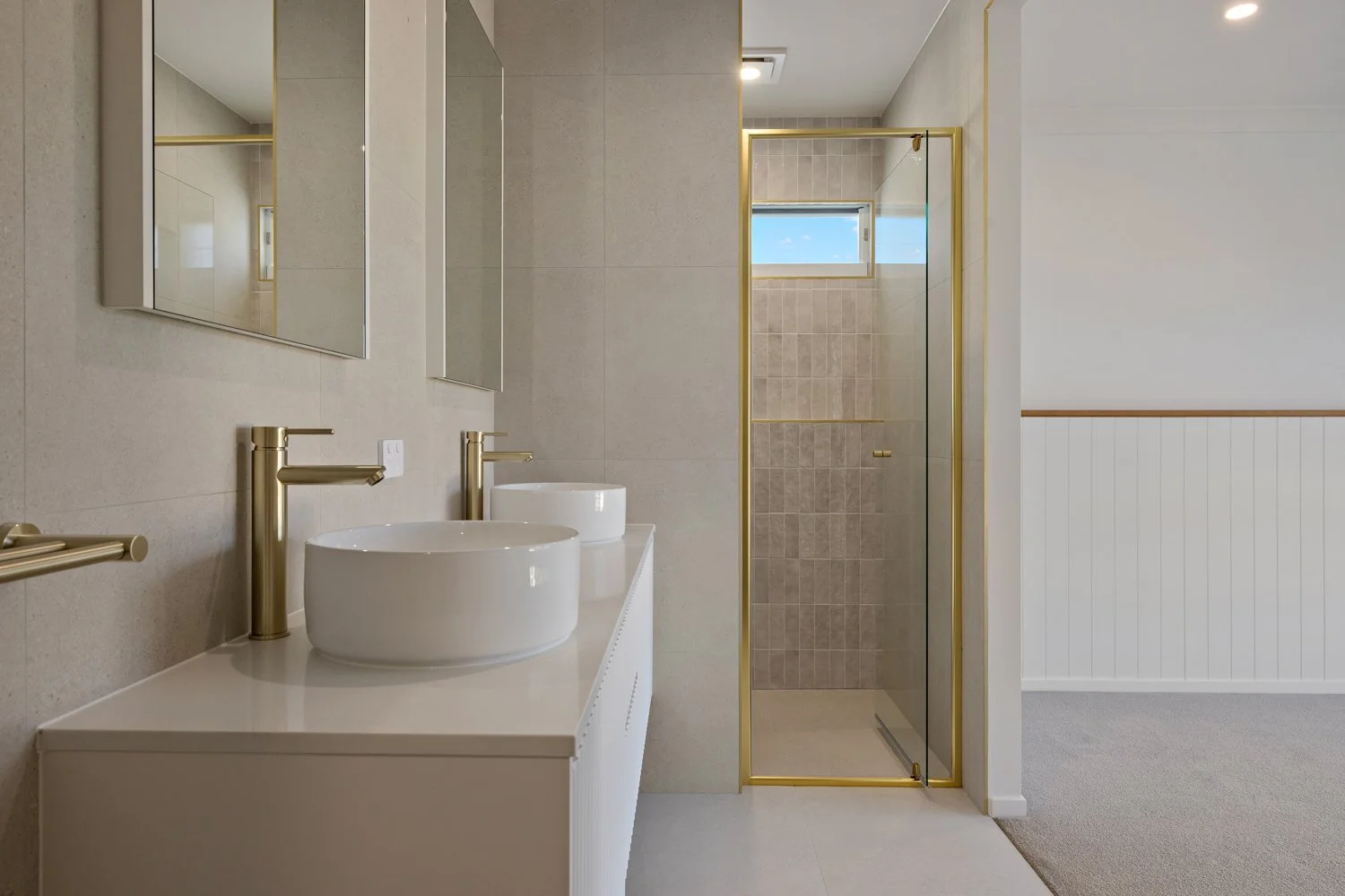Modern bathroom with beige walls and gold accents, featuring a double vanity with vessel sinks, a mirror, and a walk-in shower with glass door.