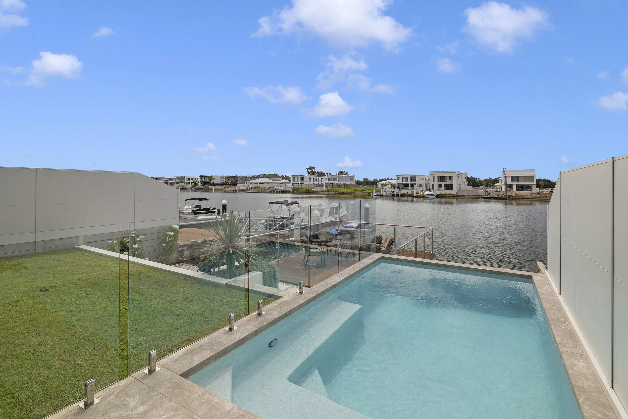 Modern backyard with a small swimming pool, glass fencing, green lawn, and a view of a river with boats and modern houses under a blue sky.