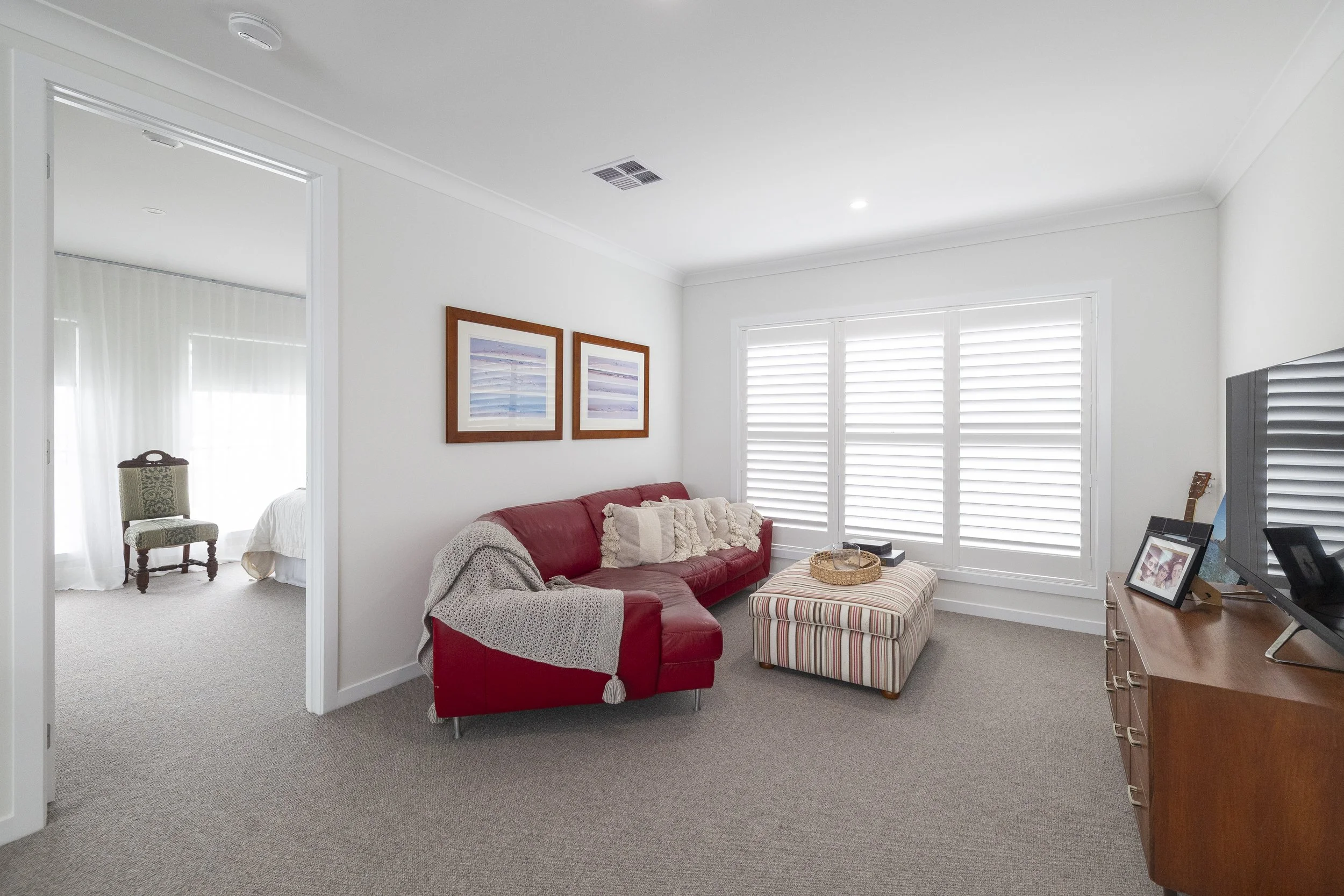 Living room with a red sofa, striped ottoman, framed artwork on white walls, and window shutters