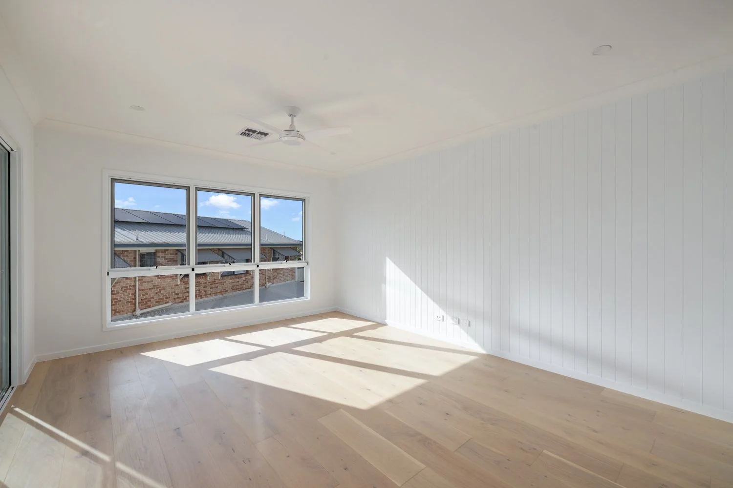 Empty room with white walls, light wood flooring, large window, ceiling fan, and sunlit view of rooftops outside.