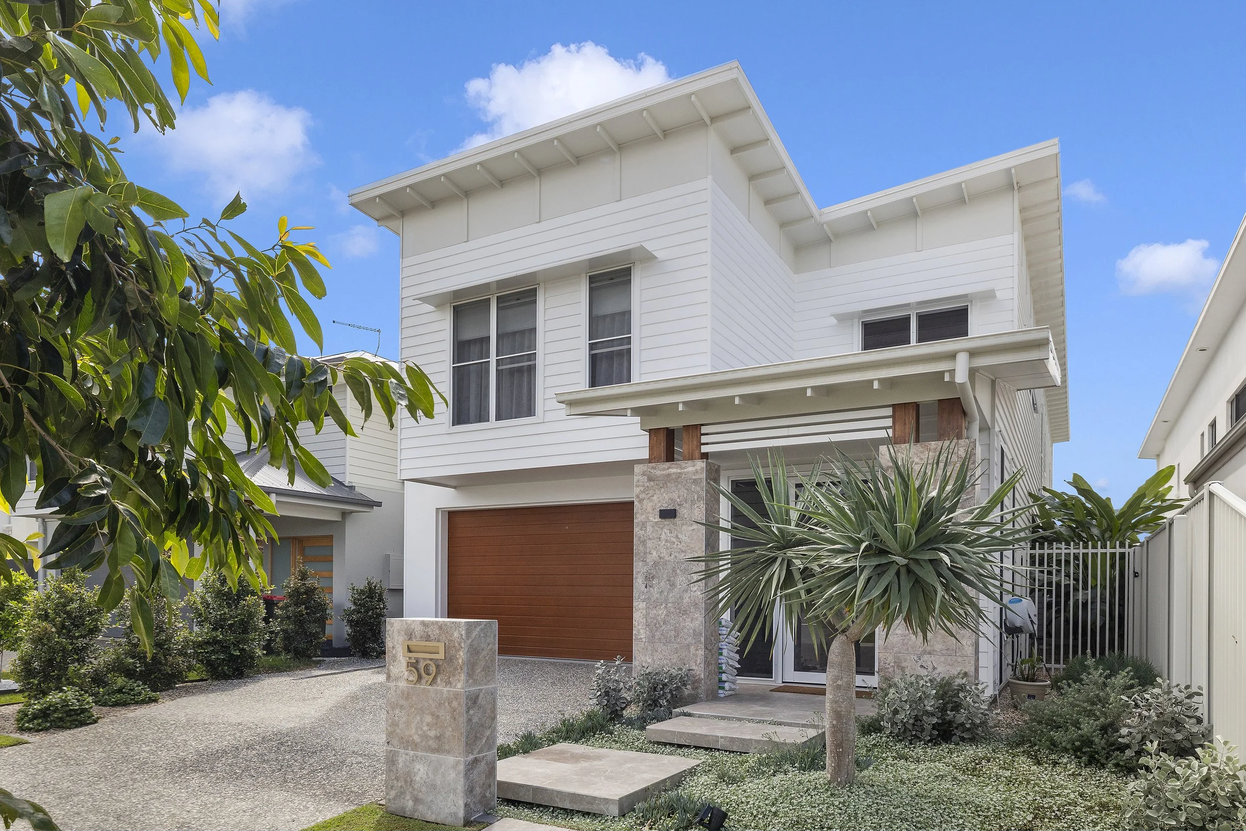 A modern white, two-story house with large windows, a wooden garage door, and a small garden with various plants and trees in the front yard.