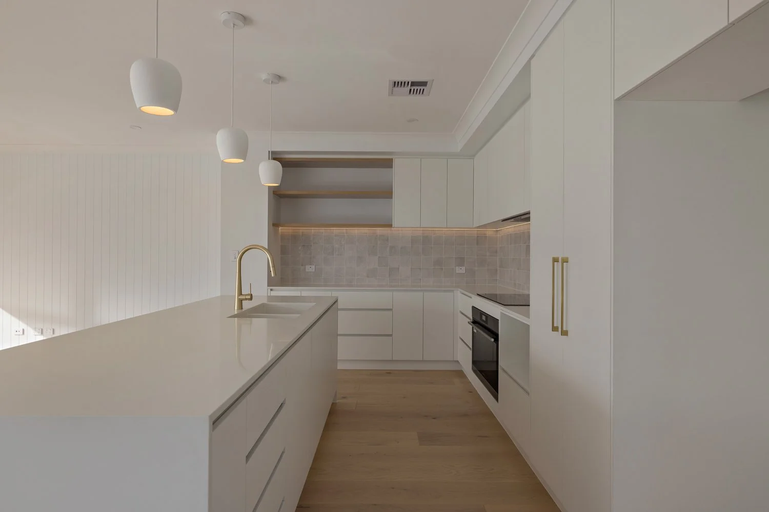 Modern kitchen with white cabinets, gold handles, built-in oven, beige tiled backsplash, white island with a gold faucet, and pendant lights.