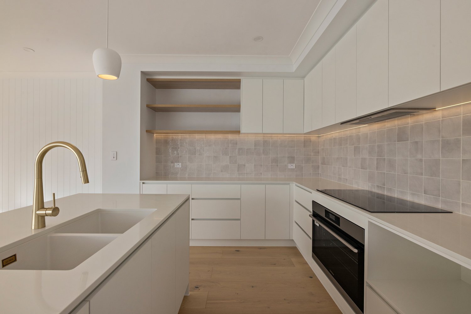 Modern kitchen with white cabinetry, beige tile backsplash, wooden open shelves, black electric stove, oven, and a white kitchen island with a gold faucet and sink.