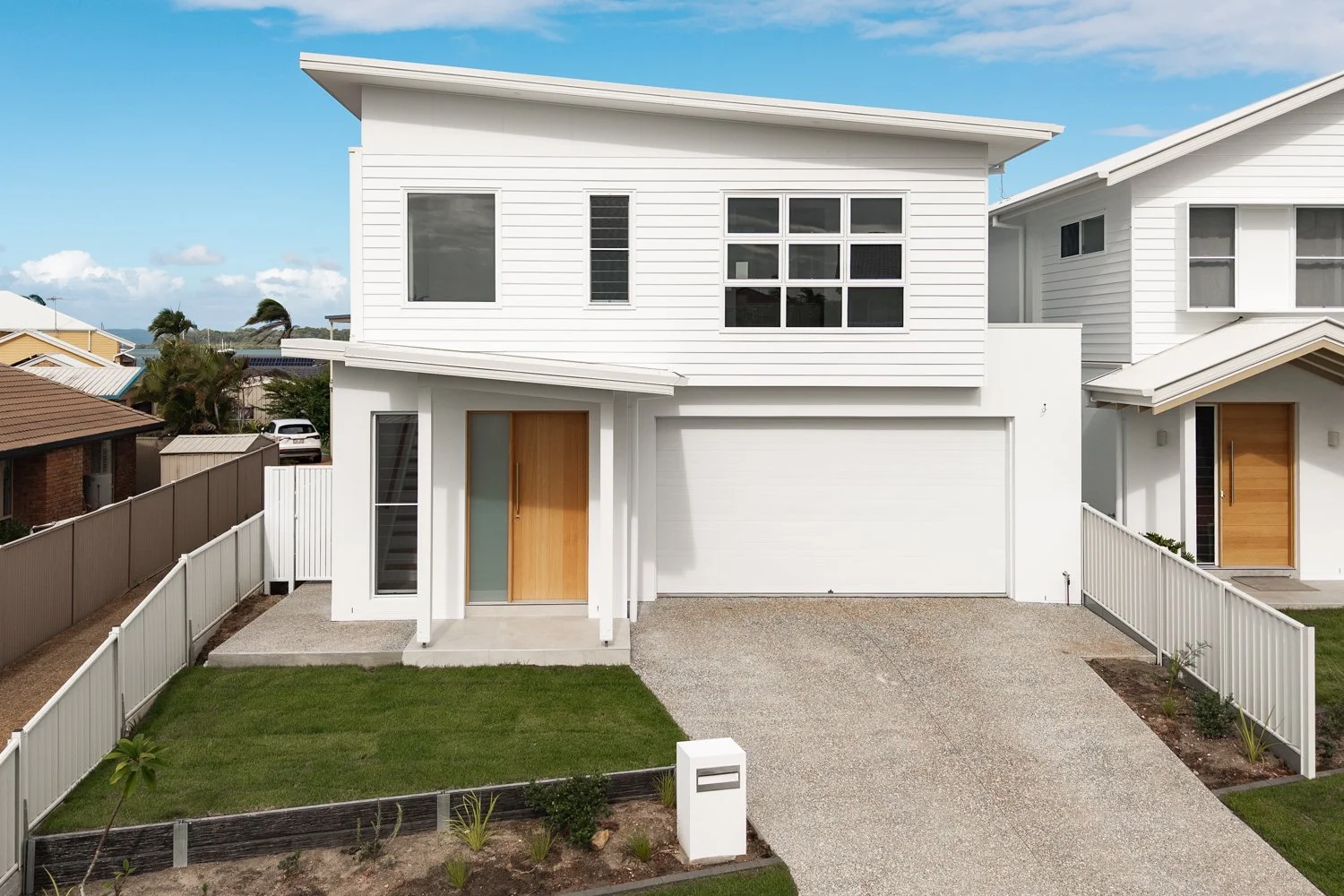 Modern two-story white house with a wooden front door, attached garage, landscaped front yard with grass and small plants, white fence, and neighboring houses under a partly cloudy sky.
