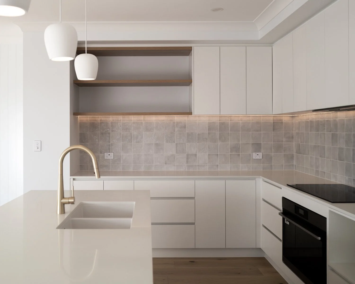 Modern kitchen with white cabinets, beige tiled backsplash, built-in oven, black stovetop, white countertop with a brass faucet and sink, and open wooden shelves on the wall.