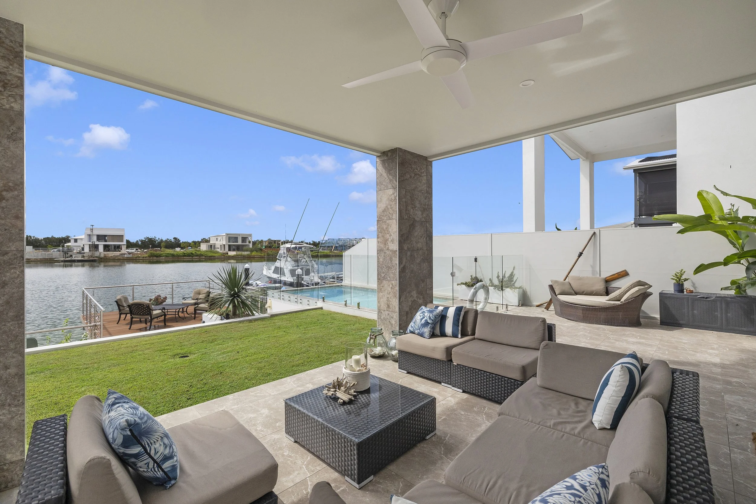 View of a covered patio area overlooking a waterway with modern houses in the background. The patio has outdoor furniture including a sofa, chairs, a black wicker coffee table, and decorative cushions. There is a ceiling fan on the ceiling.