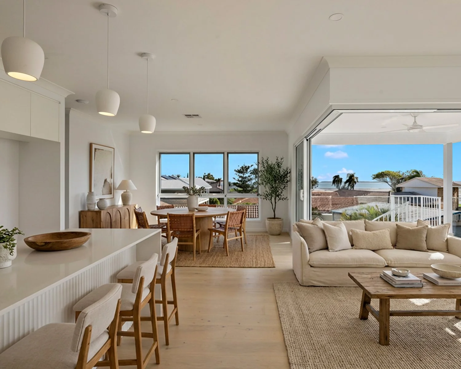 Living room with a beige sofa, wooden coffee table, dining area with a round table and four chairs, large sliding glass doors leading to a balcony, and ocean views with palm trees in the distance.