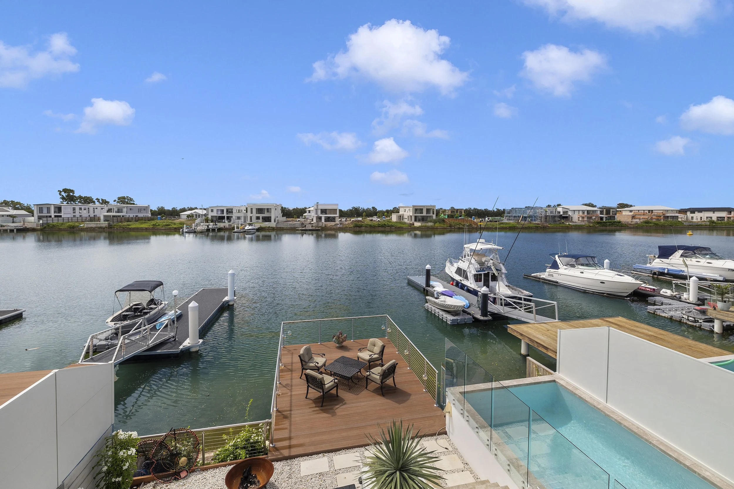 Panoramic view of a marina with yachts docked along the water, modern houses in the background, blue sky with clouds, and outdoor seating on a wooden deck.