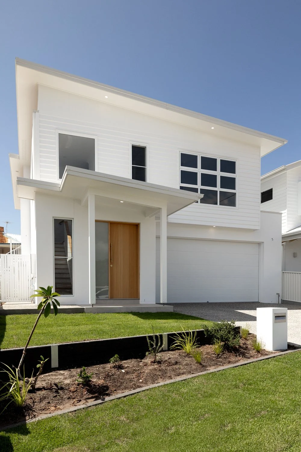 Modern white two-story house with large windows, a wooden front door, and a well-maintained lawn with small plants in the garden bed.