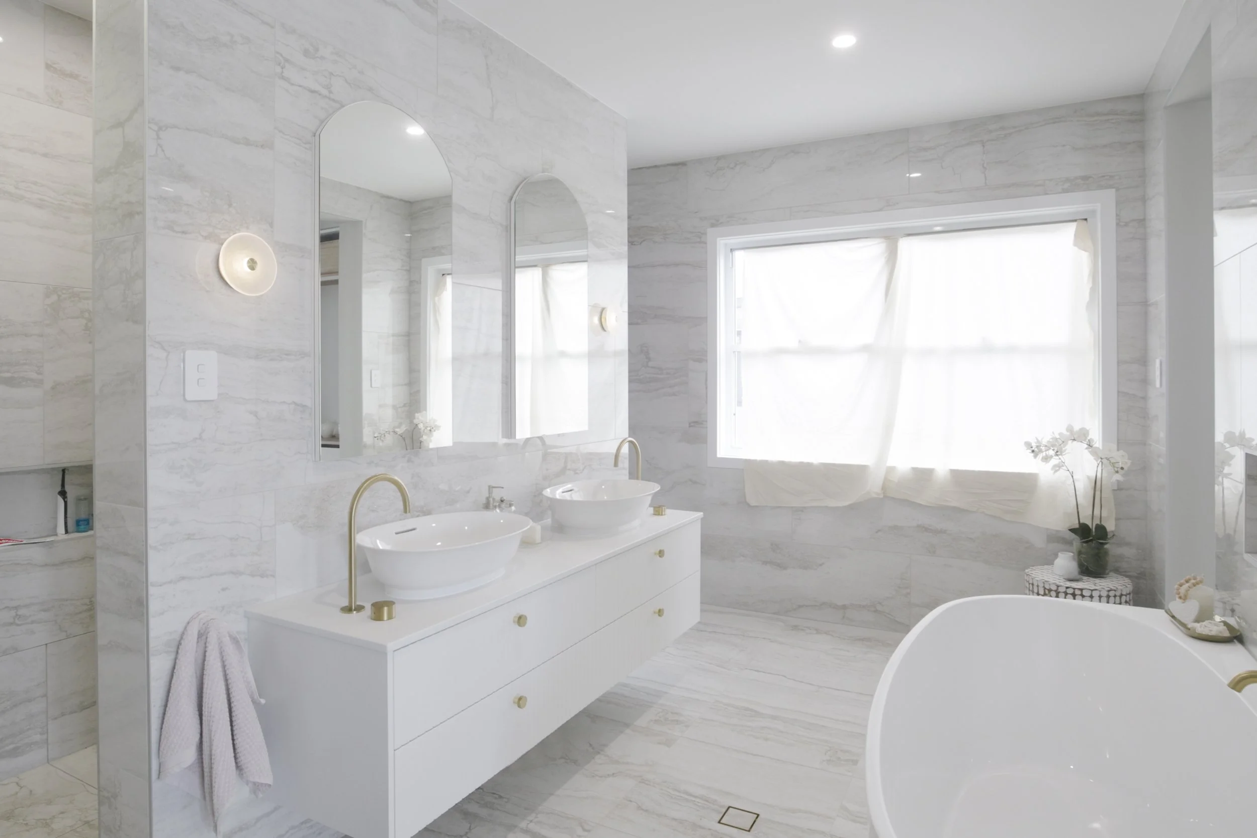 A modern, bright bathroom with white marble walls and floors, a double sink vanity, oval mirrors, a large window with white curtains, and a bathtub with decorative plants and toiletries.