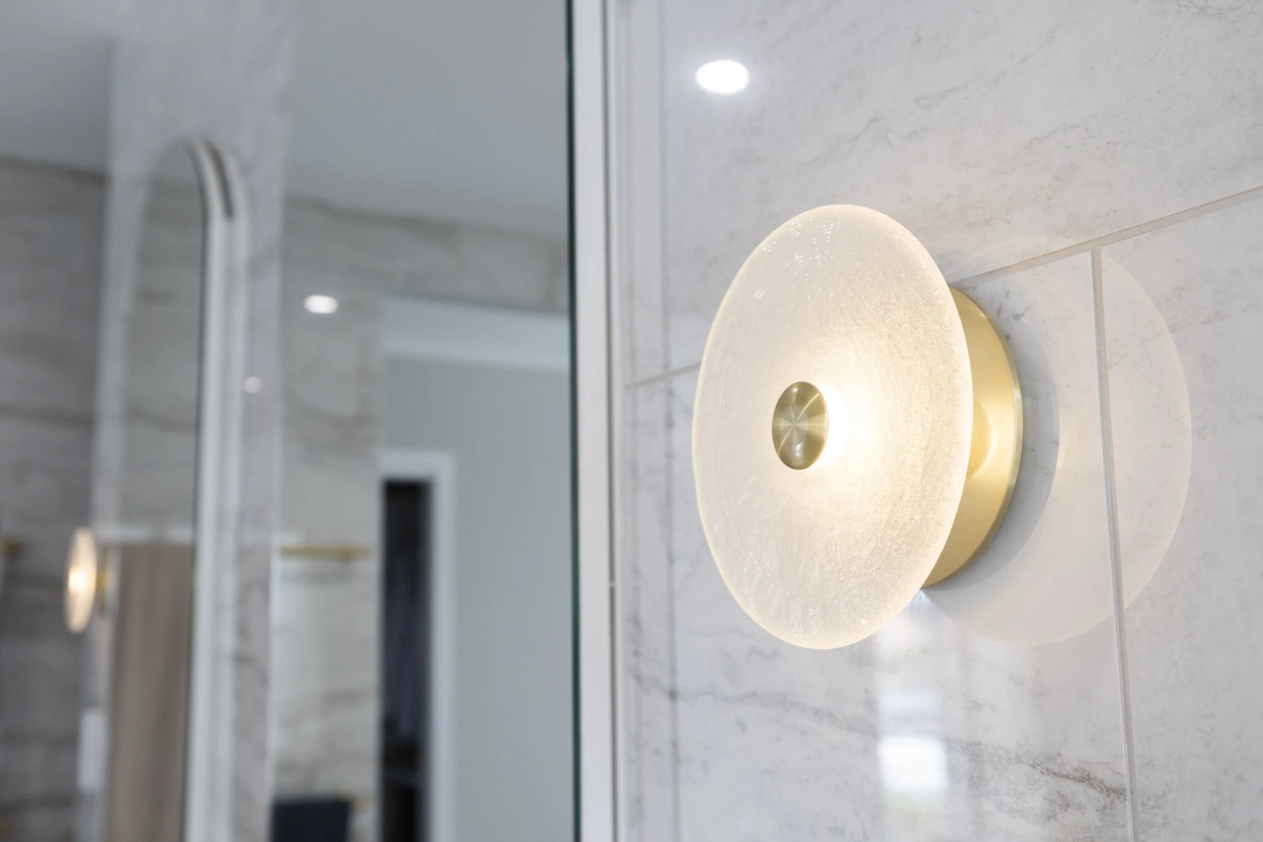 Close-up of a round wall-mounted lamp with a white, textured surface and a metallic center, illuminating a light-colored tiled wall.