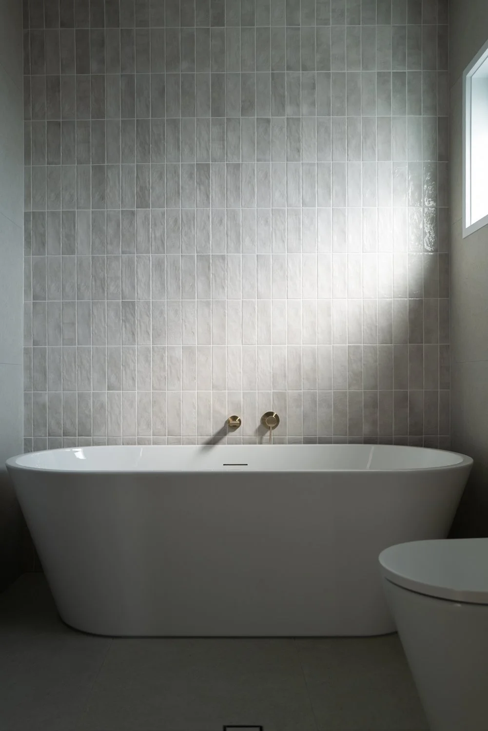 Modern white bathtub with gold fixtures against a tiled wall in a minimalist bathroom with a window.